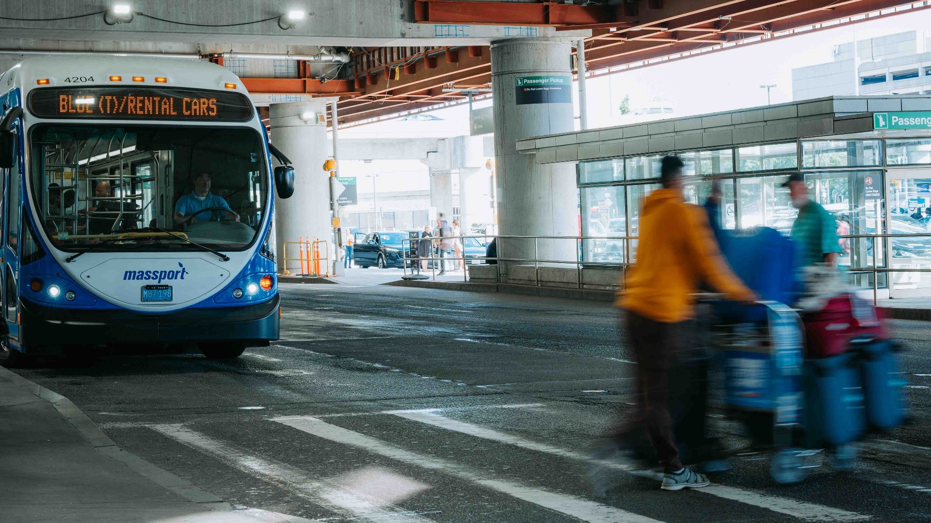 Busy airport scene showing travelers in motion and a rental car bus waiting under a covered terminal.
