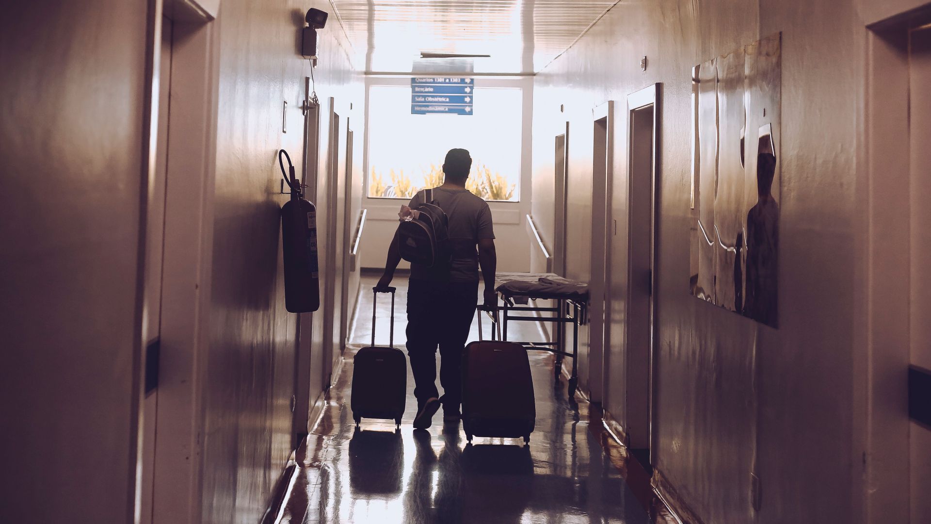 Silhouette of a traveler with luggage walking down an illuminated hotel corridor.