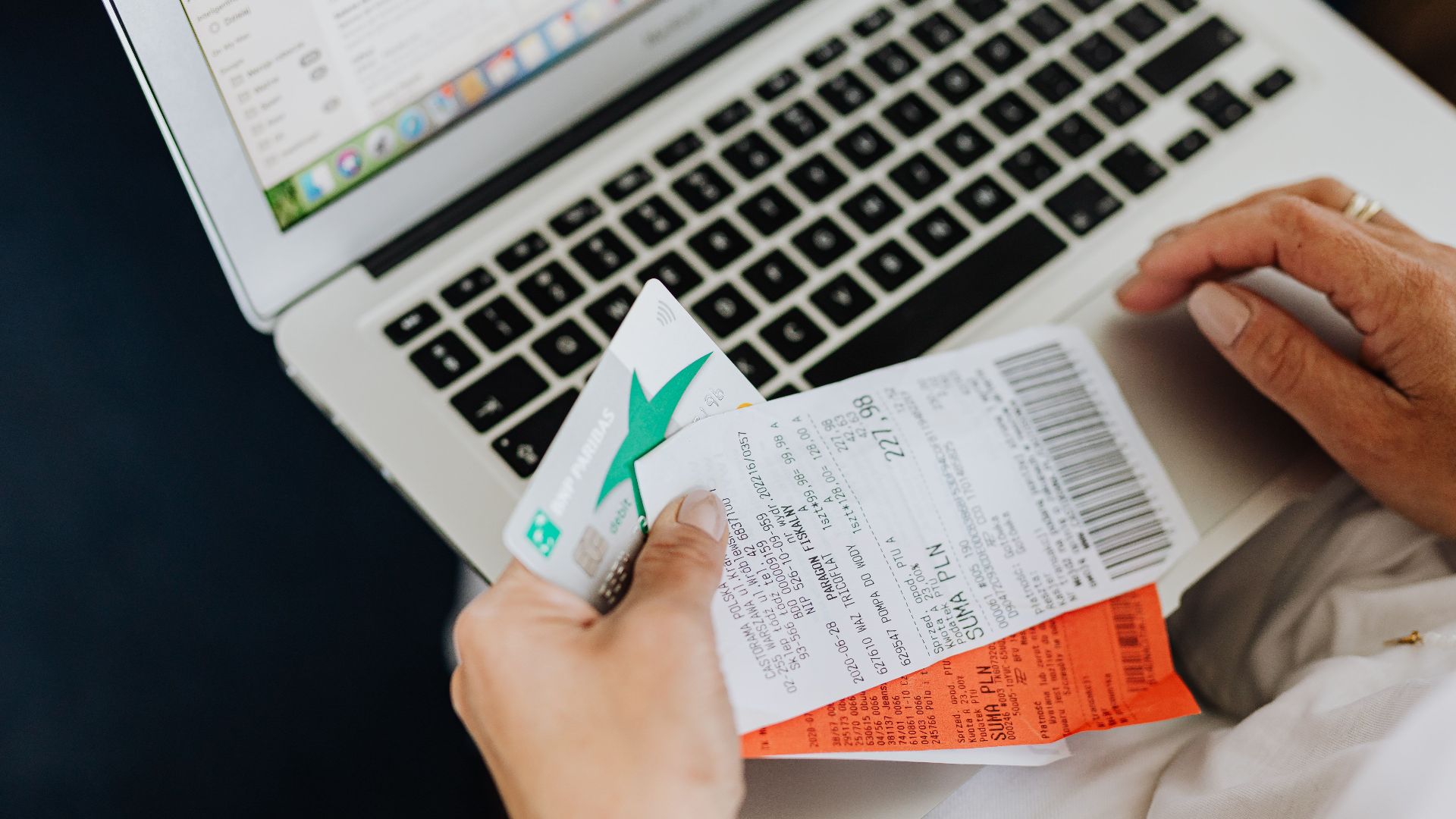 Close-up of hands holding receipts and a bank card in front of a laptop, representing online shopping and e-commerce.