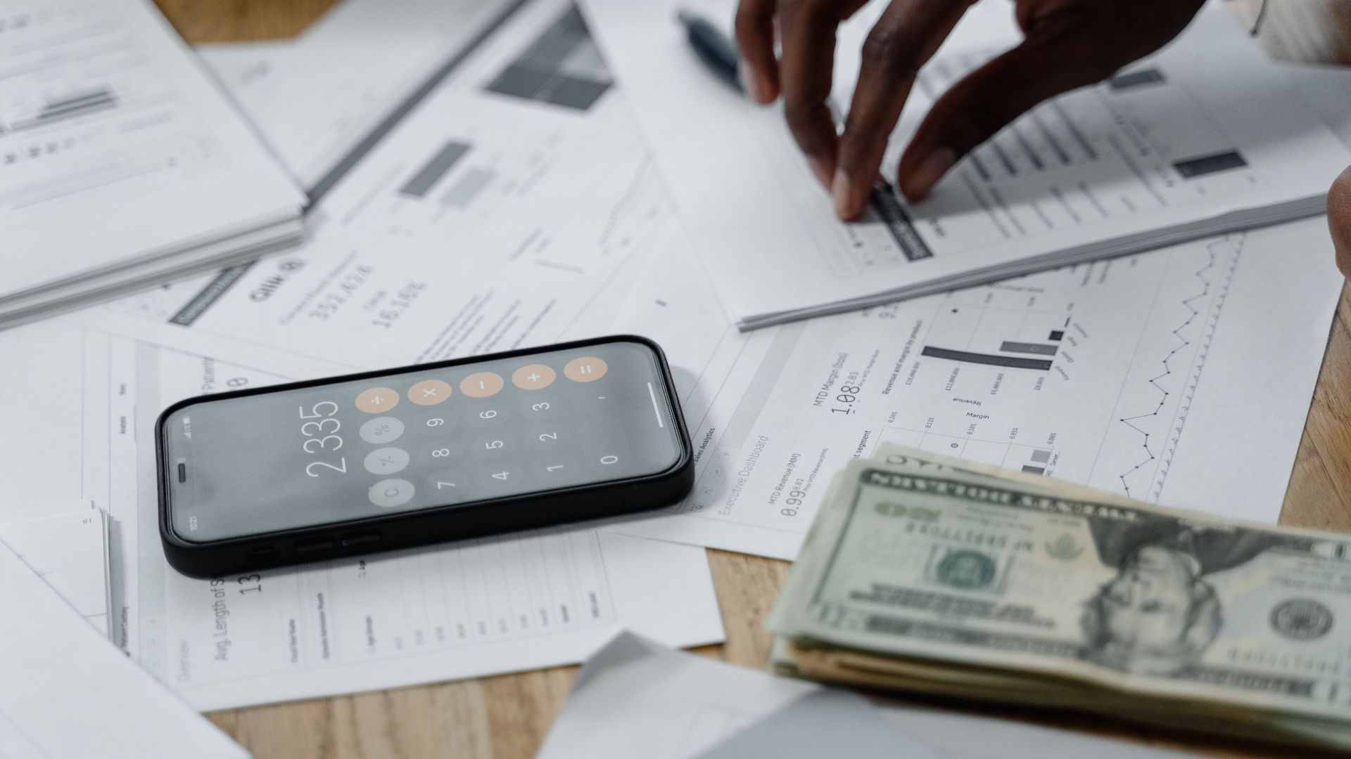 Close-up of hands calculating financial data with reports, smartphone, and cash on table.