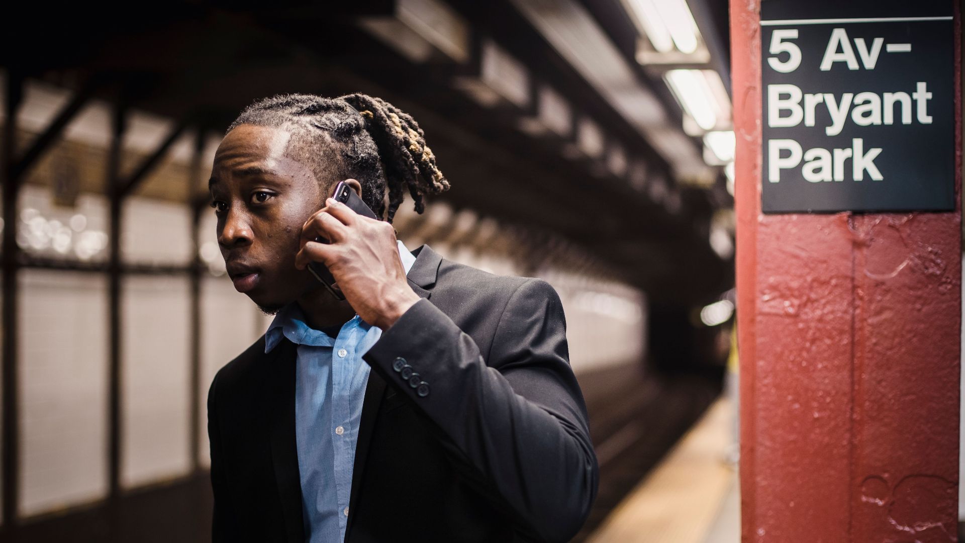 African American male in formal wear talking on mobile phone and looking into subway tunnel with worried expression on face while waiting for train at empty New York metro station