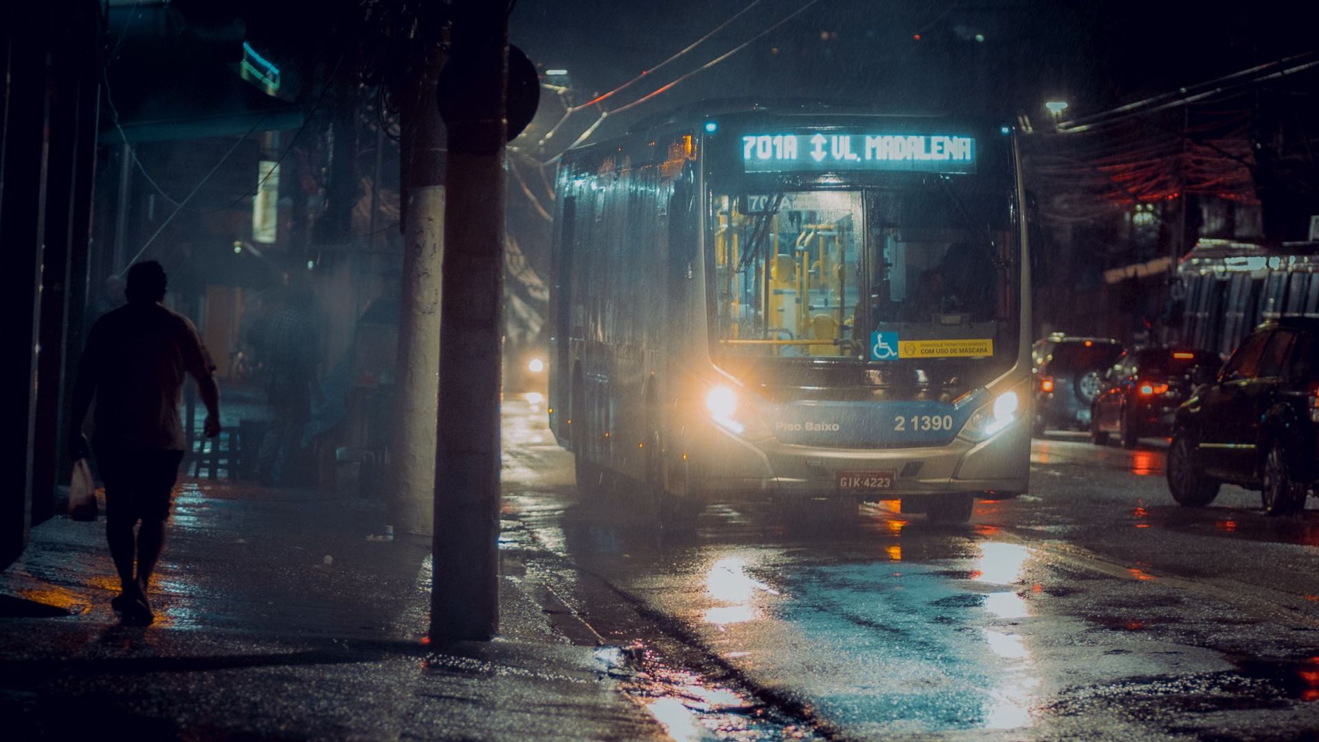 A city bus travels a wet urban street at night, reflecting city lights and atmosphere.