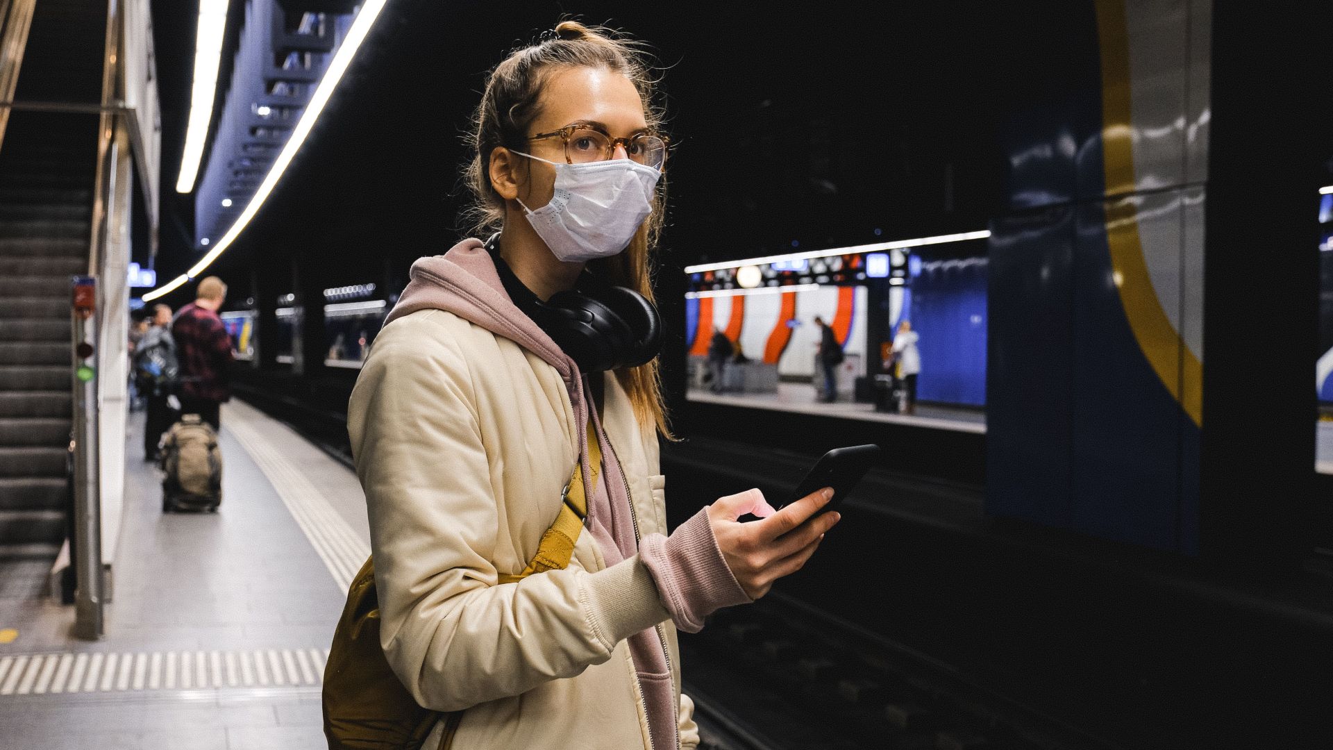 Woman with face mask holding smartphone in urban subway station during pandemic.