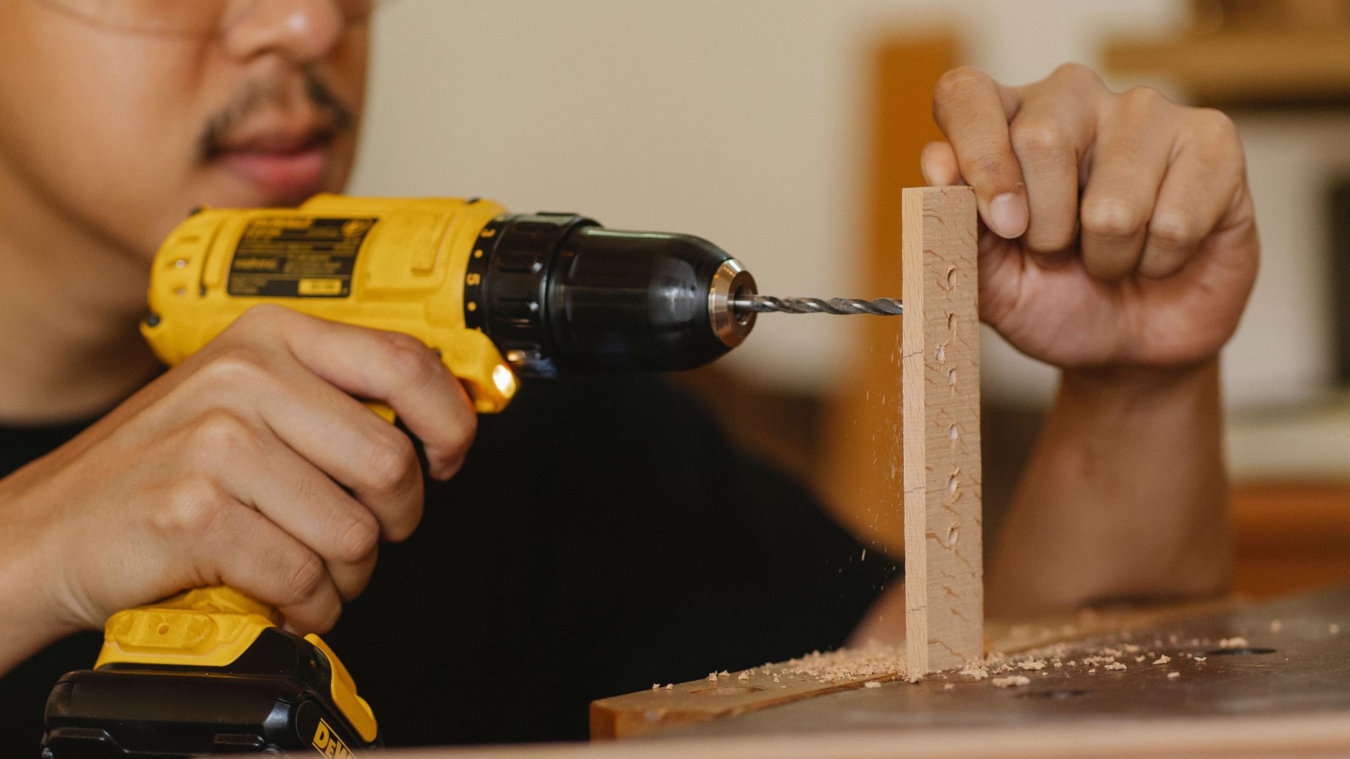 Crop anonymous male drilling holes in wooden panel while working on table in workshop