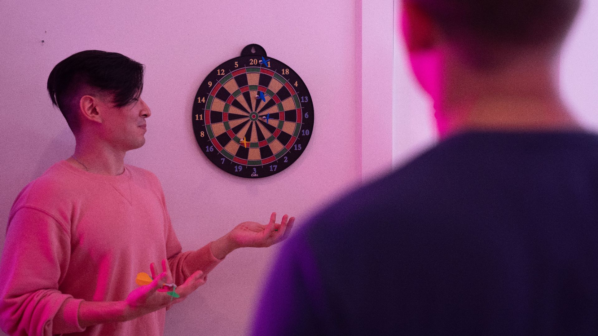 Two young men playing darts indoors with vibrant pink lighting, focusing on the dartboard.