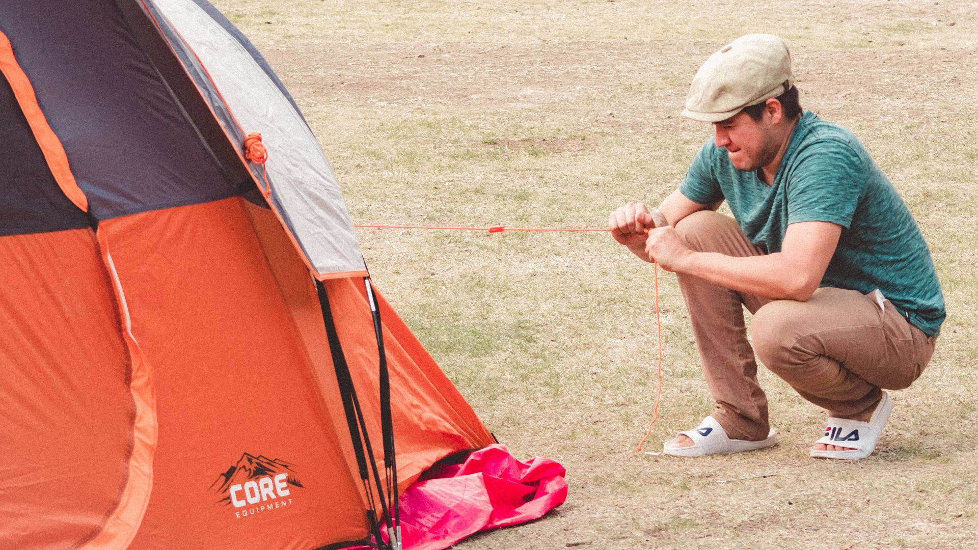A man in casual attire sets up an orange tent at a campground during the day, showing a relaxed outdoor activity.