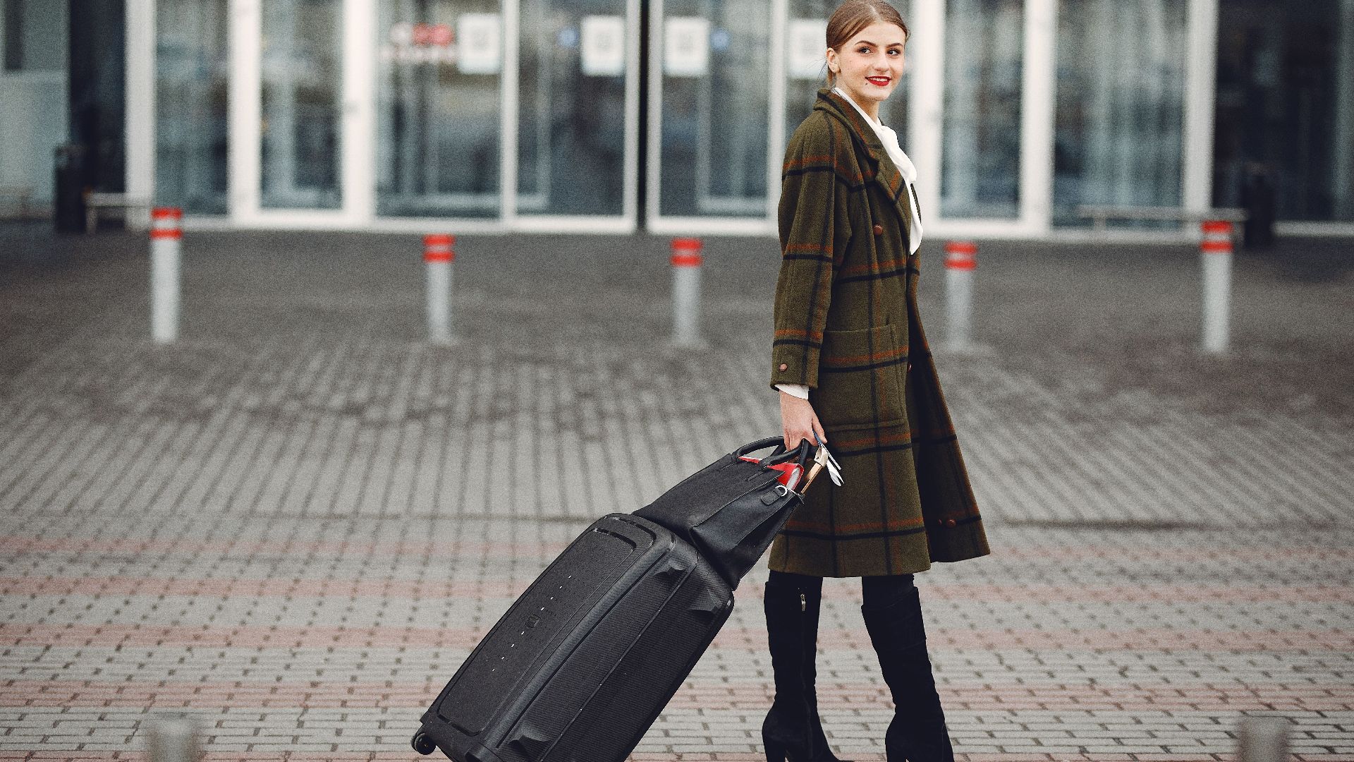 Full length of smiling female manager in stylish coat walking with suitcase and bag while traveling on business trip smiling away