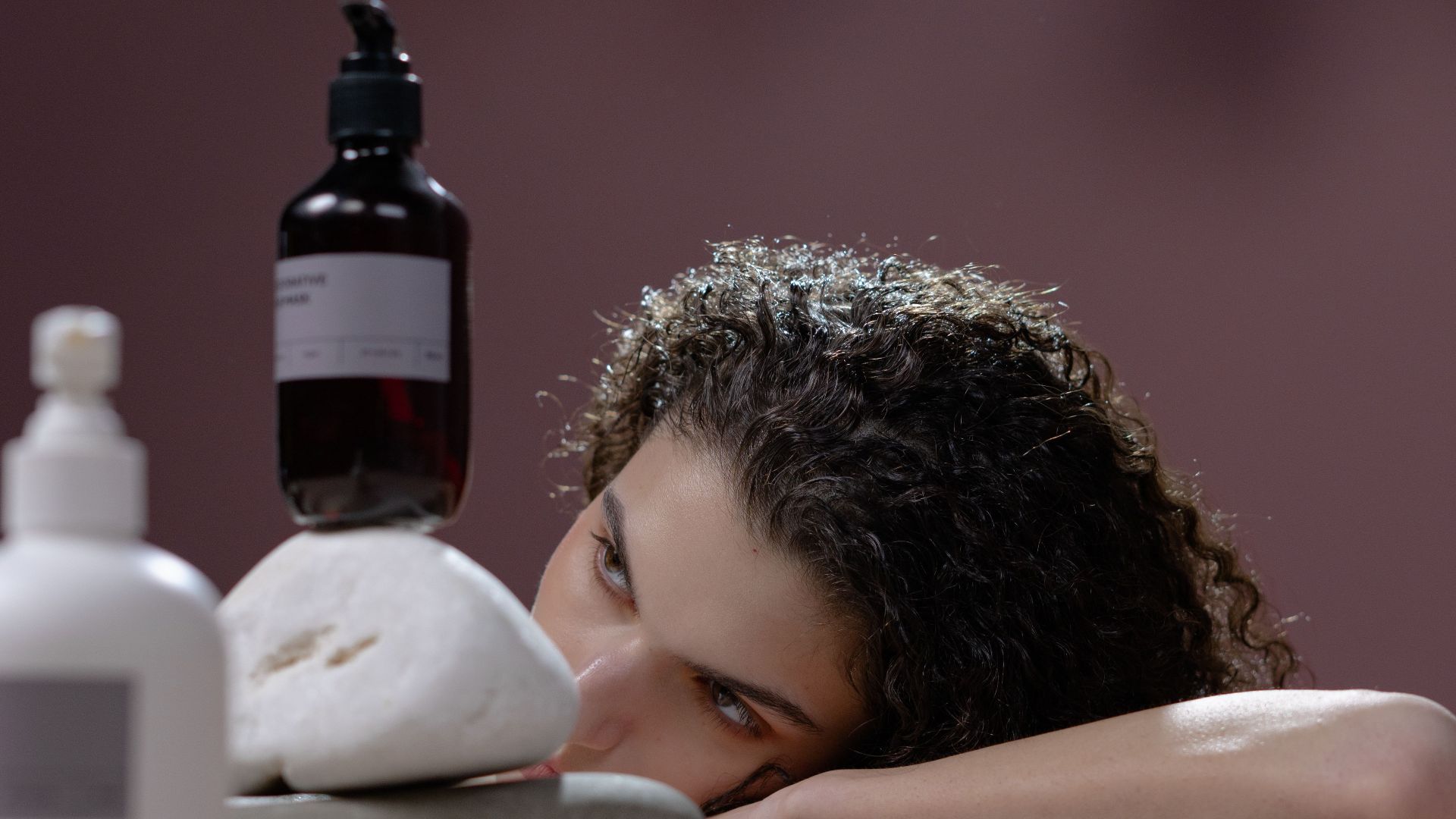 A woman with curly hair leans on a table holding cosmetic bottles atop stacked stones.