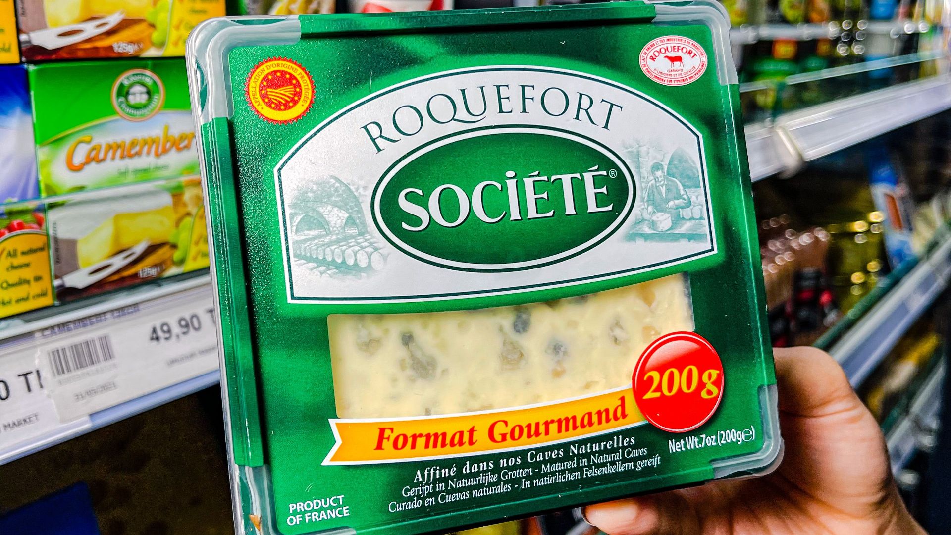 Close-up of a hand holding Roquefort cheese in a supermarket cheese aisle.