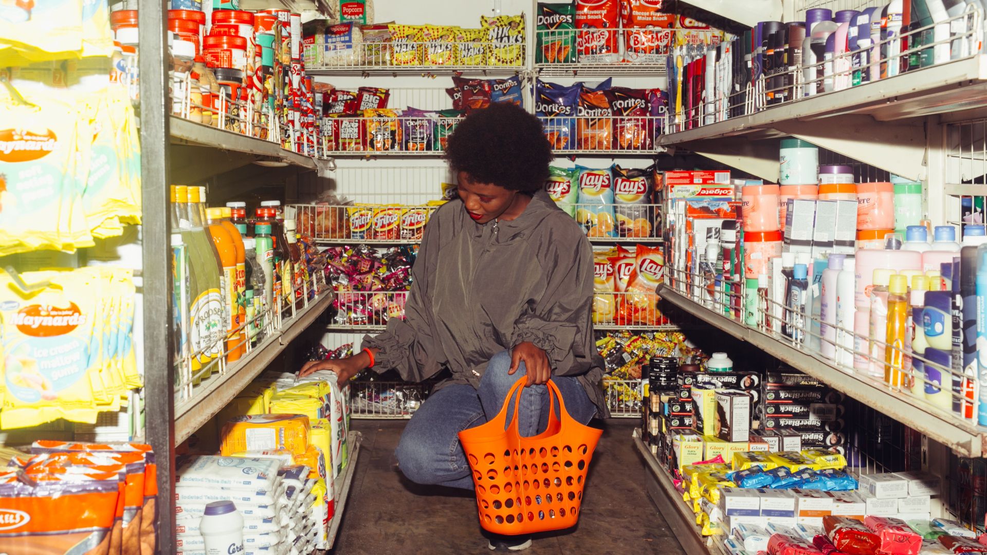 Full length of adult ethnic female buyer selecting goods while squatting with shopping bag among grocery shelves