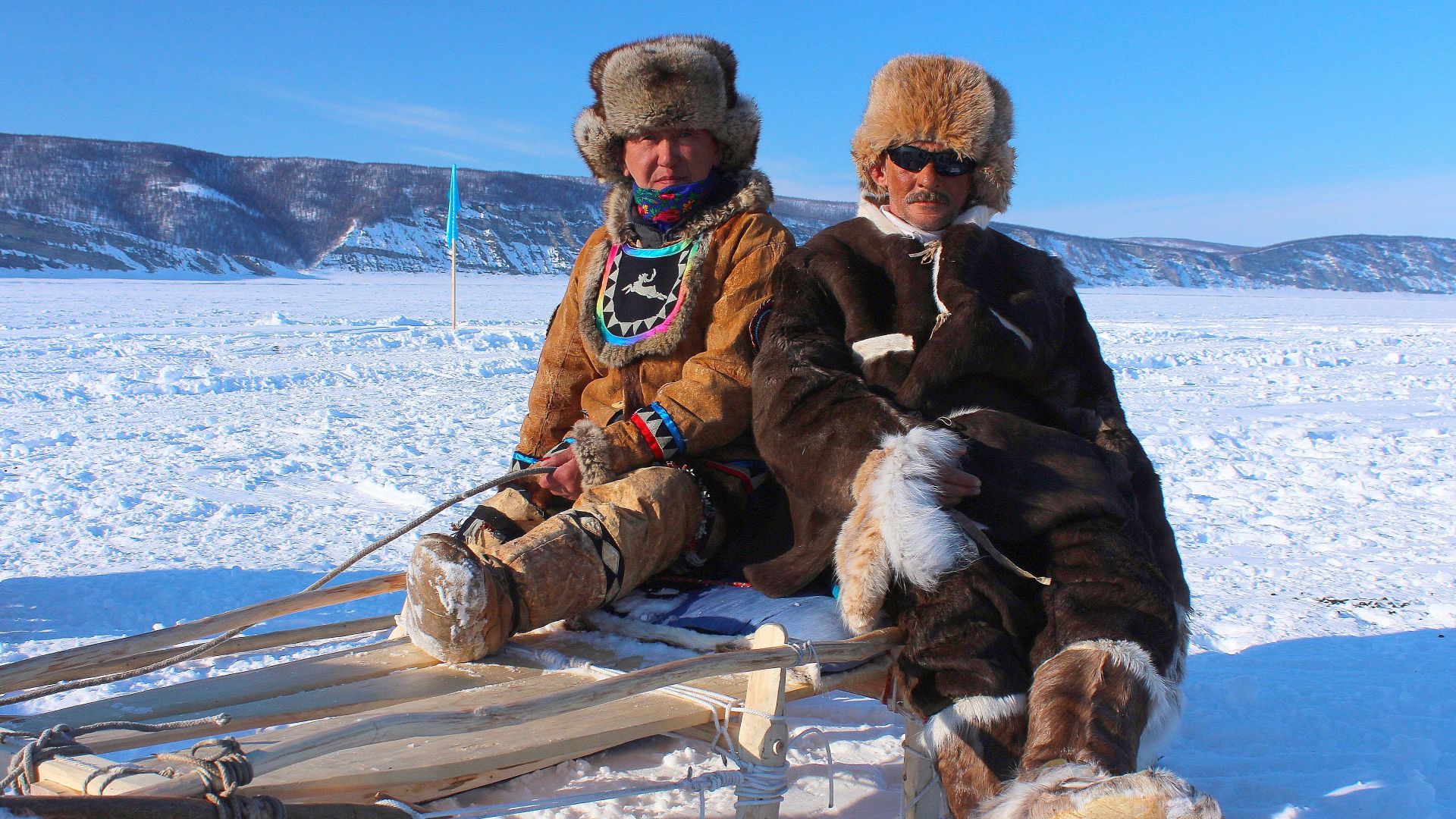 Two adults in traditional fur clothing sitting on a wooden sled in snowy Siberia.