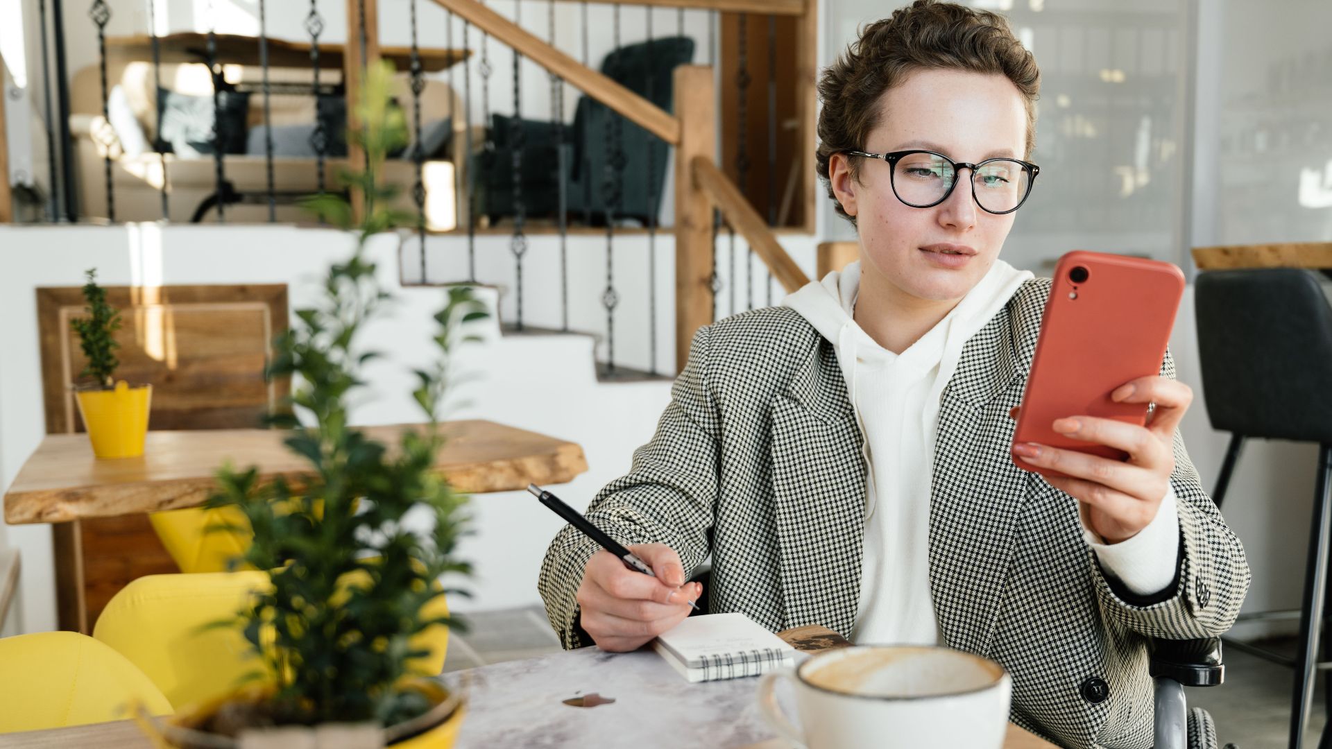 Concentrated businesswoman in formal wear and eyeglasses using wheelchair browsing smartphone and taking notes in copybook while working in cafe