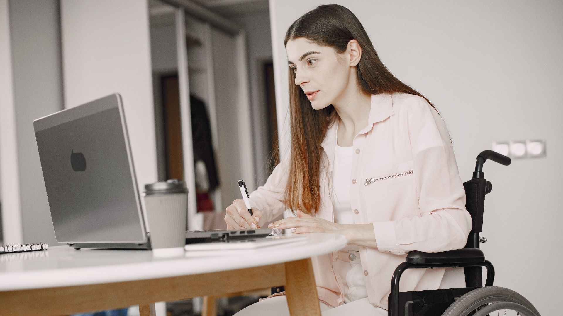 Woman in a wheelchair working remotely at home with a laptop and taking notes, showcasing remote work and accessibility.