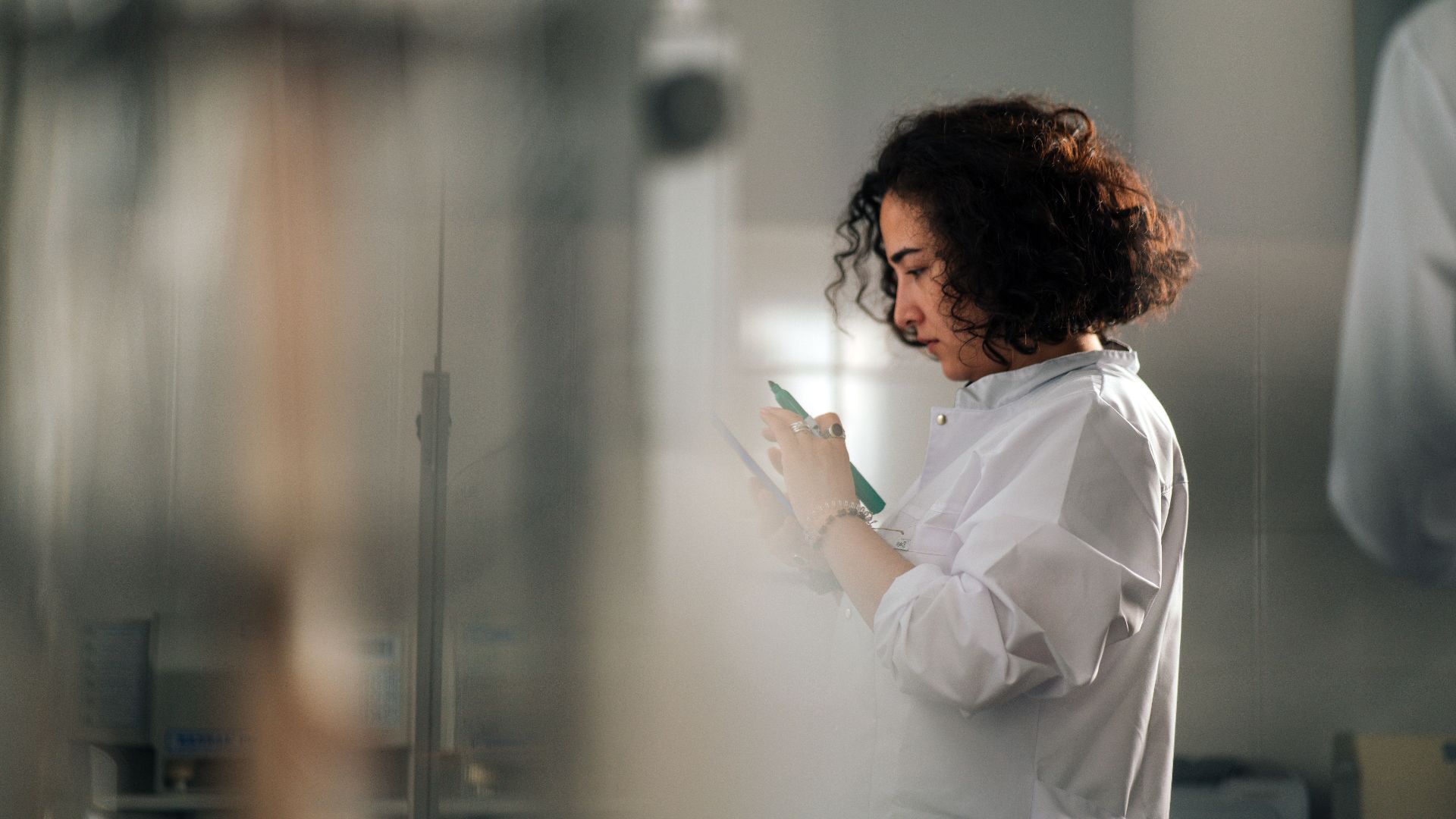A focused female scientist with curly hair writes notes in a laboratory setting.