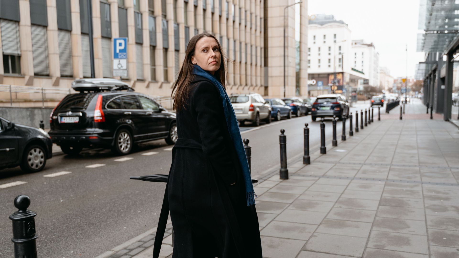 Professional woman in black coat walking on urban sidewalk during the day.