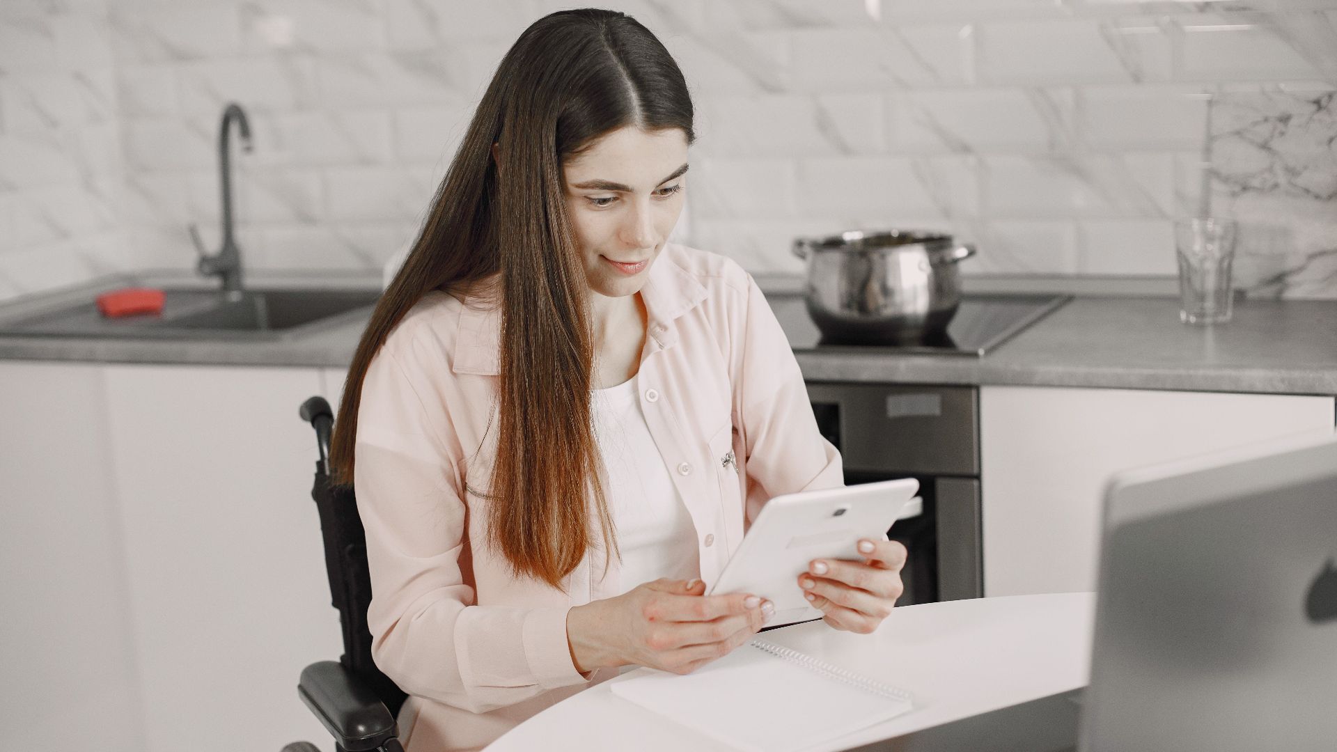 Woman in wheelchair engaged with tablet at kitchen table, creating an inclusive work environment.