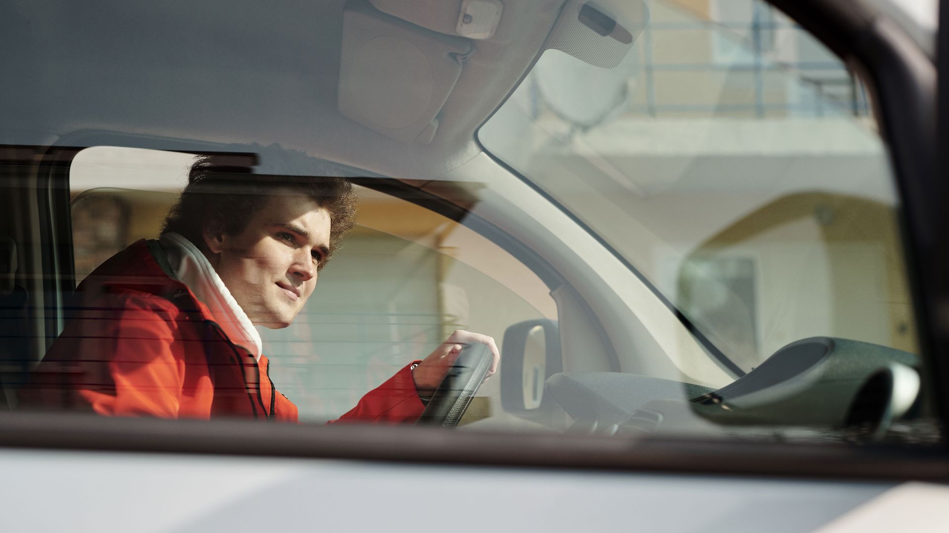 A man in a red jacket driving a car, seen through the window on a sunny day.