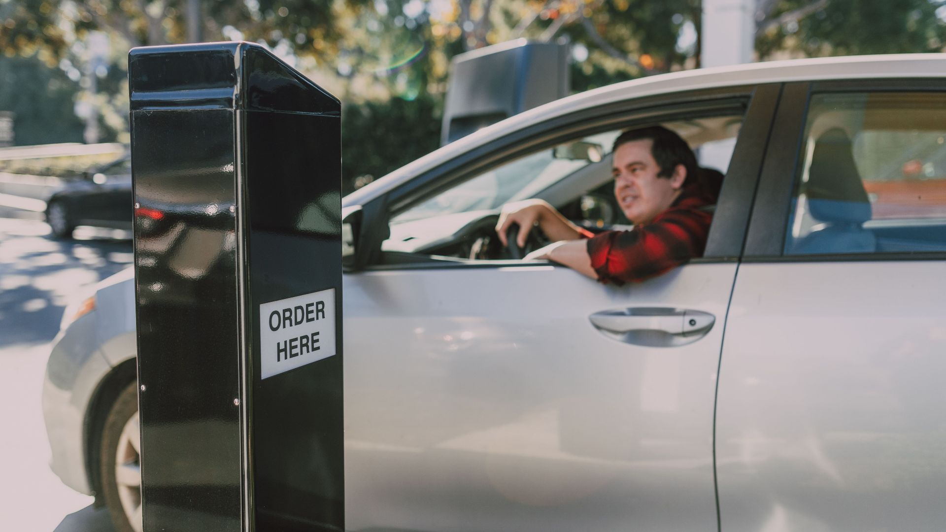 A man in a car orders at a drive-thru window, surrounded by outdoor scenery.