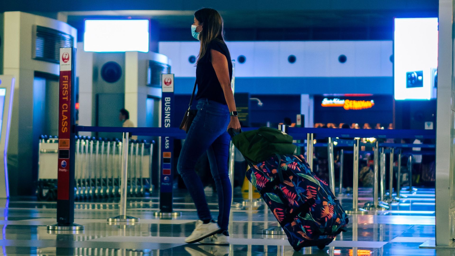 A woman wearing a face mask walks through an airport with luggage, reflecting modern travel trends.