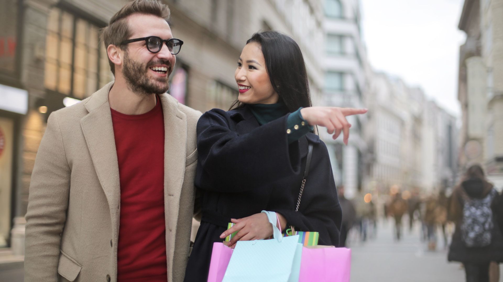 From below happy stylish Asian woman in warm clothes smiling at cheerful unshaven man in glasses and pointing finger away while walking along street together in city after shopping