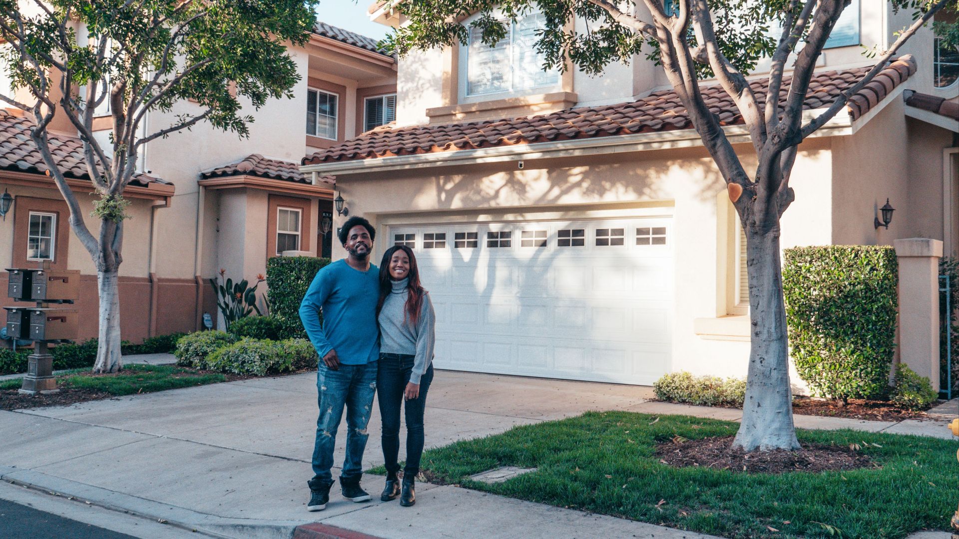 A smiling couple proudly posing in front of their newly purchased house, expressing joy and togetherness.