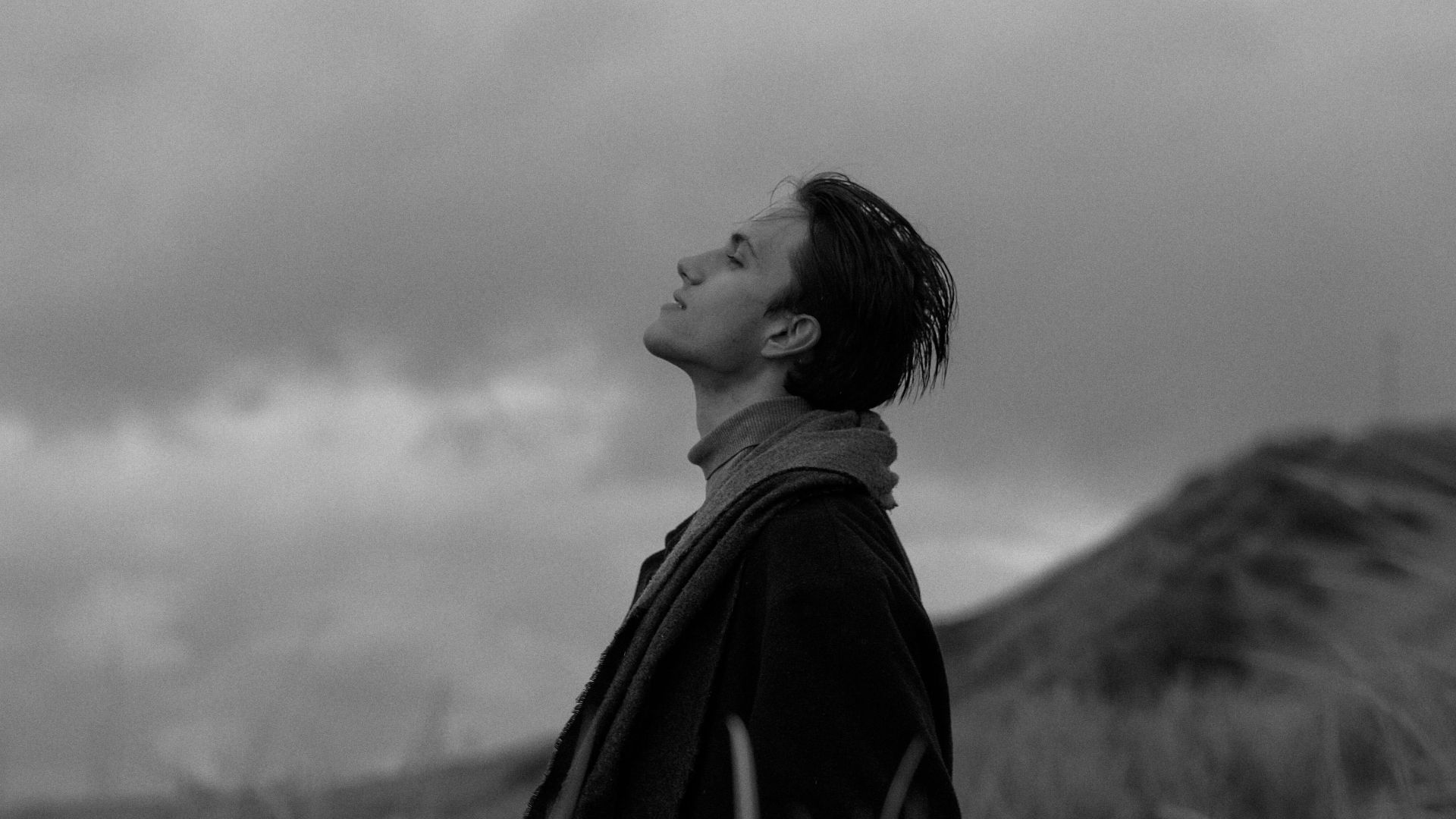 Black and white portrait of a man in a coat, standing in a grassy field under a cloudy sky.
