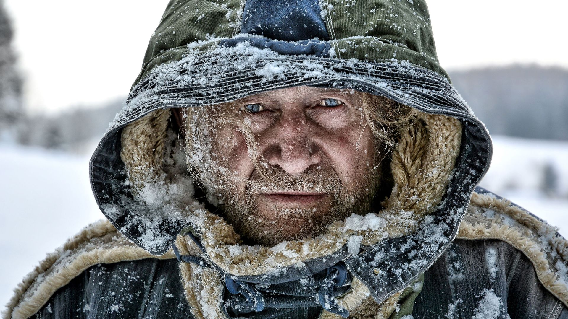 Close-up portrait of a mature man in a hooded jacket covered in snow, showcasing the harsh winter weather.