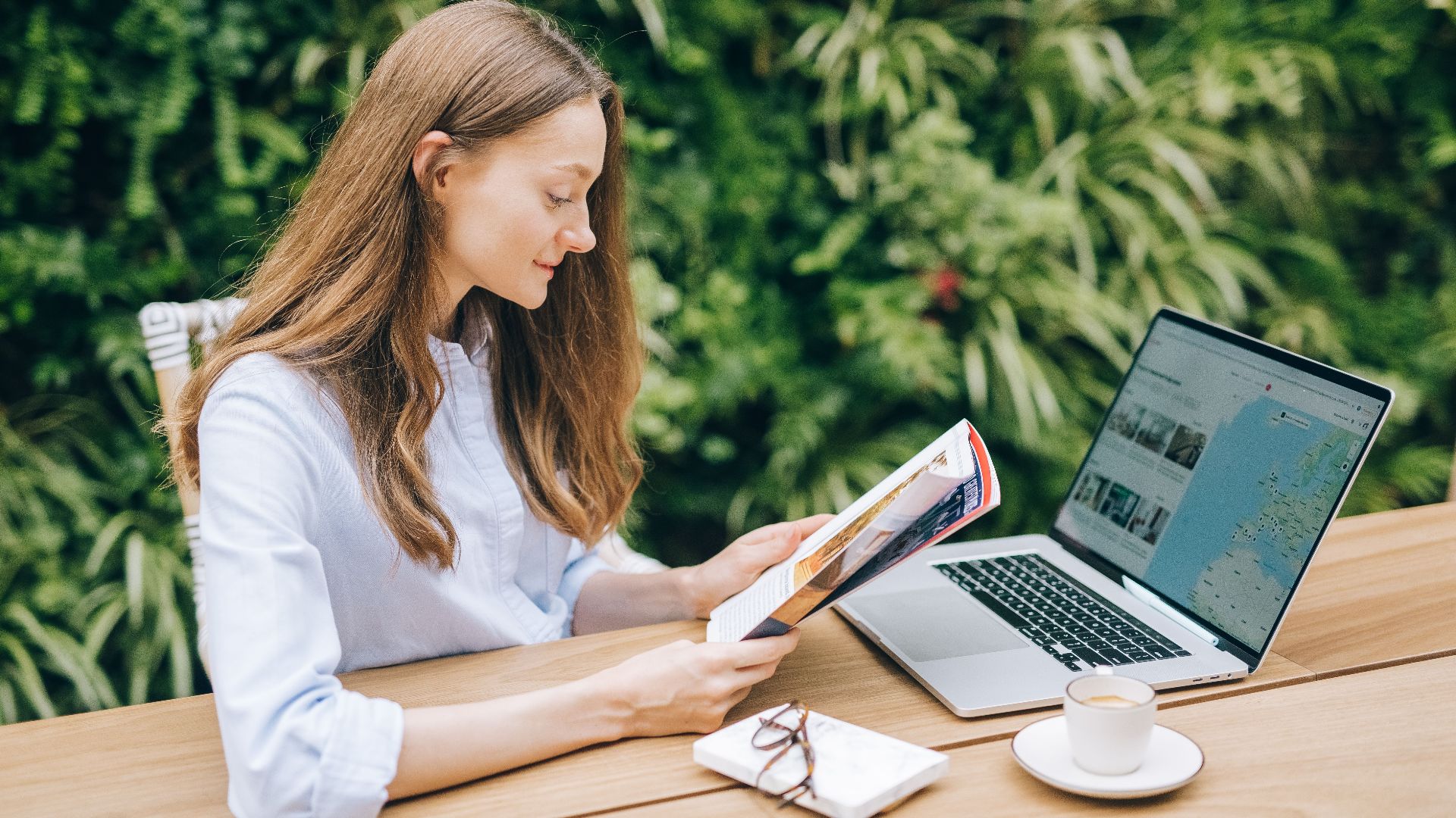 Caucasian woman reading a magazine at a wooden table with a laptop, outdoors.