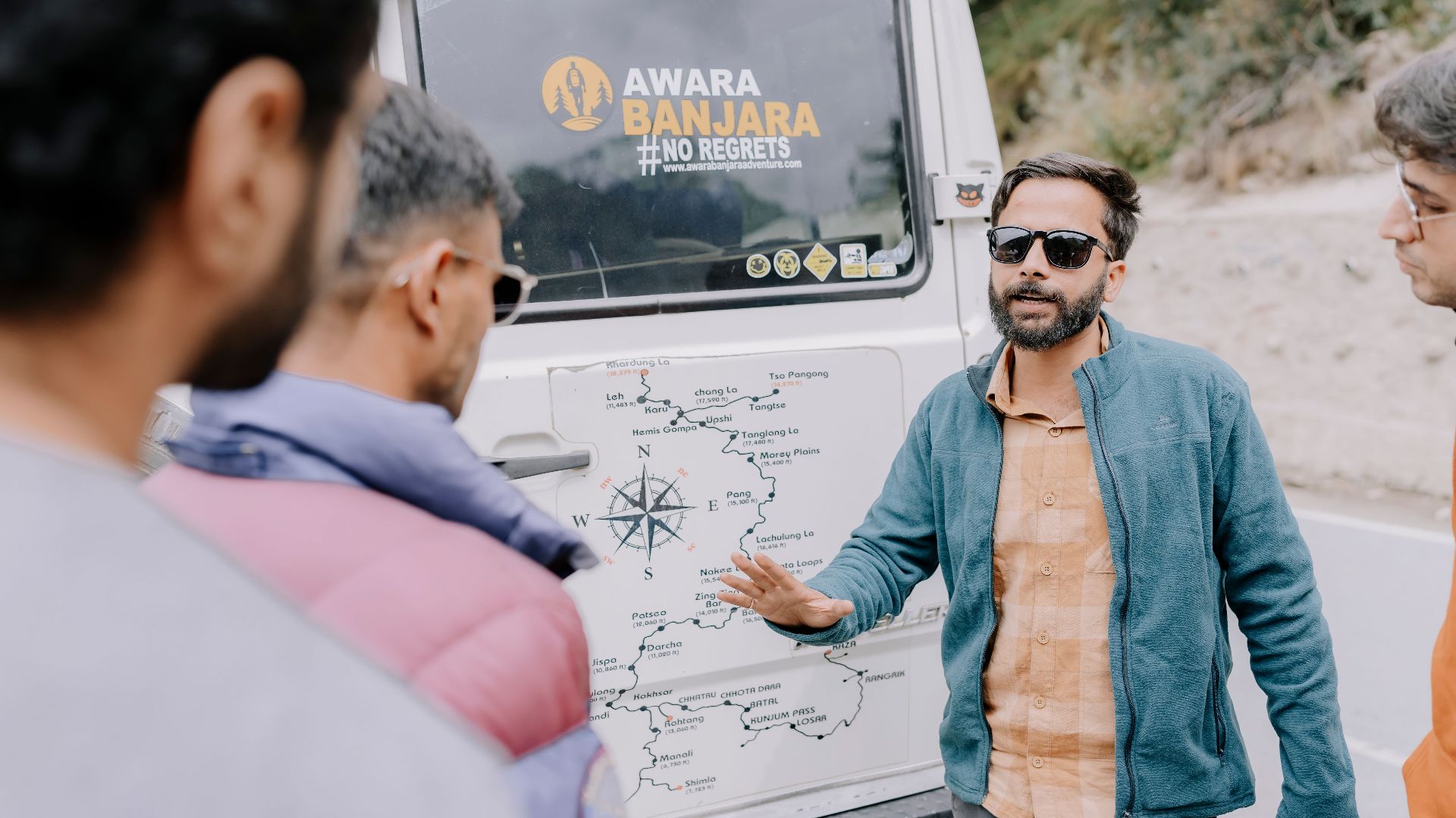 A guide in a blue jacket explains a travel route to three adults near a vehicle