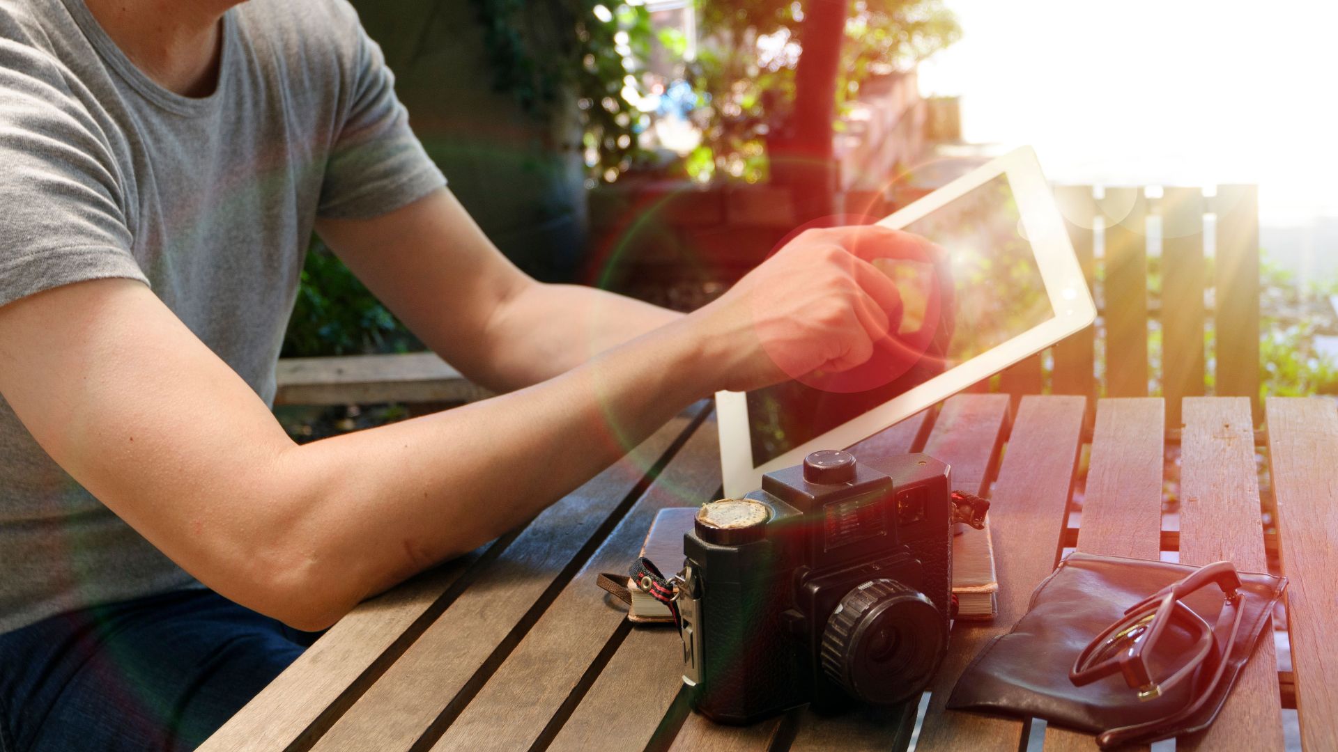 A person uses a tablet outdoors with a vintage camera and sunglasses nearby.