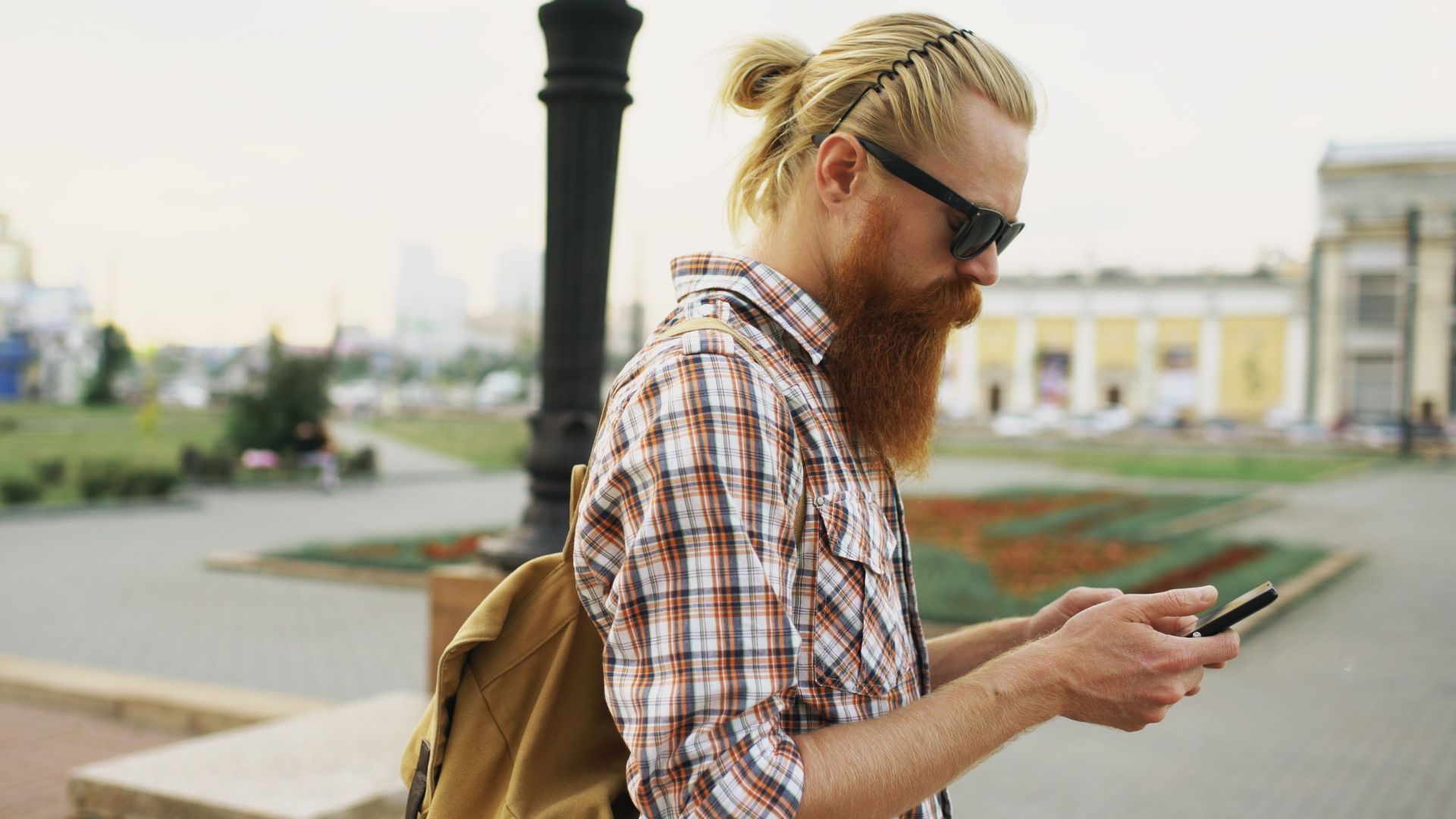 Bearded man in sunglasses and plaid shirt using smartphone outdoors.