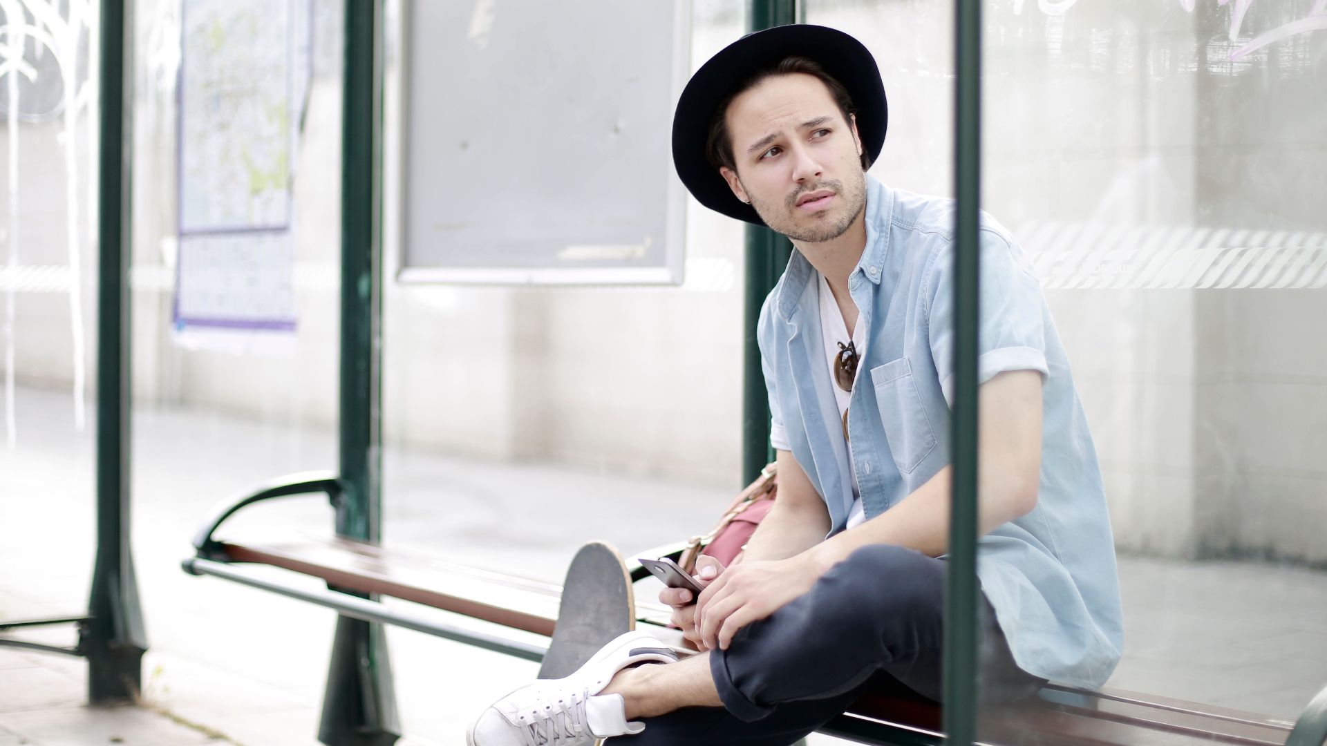 Stylish man in hat and sunglasses sitting with a skateboard at a bus stop.