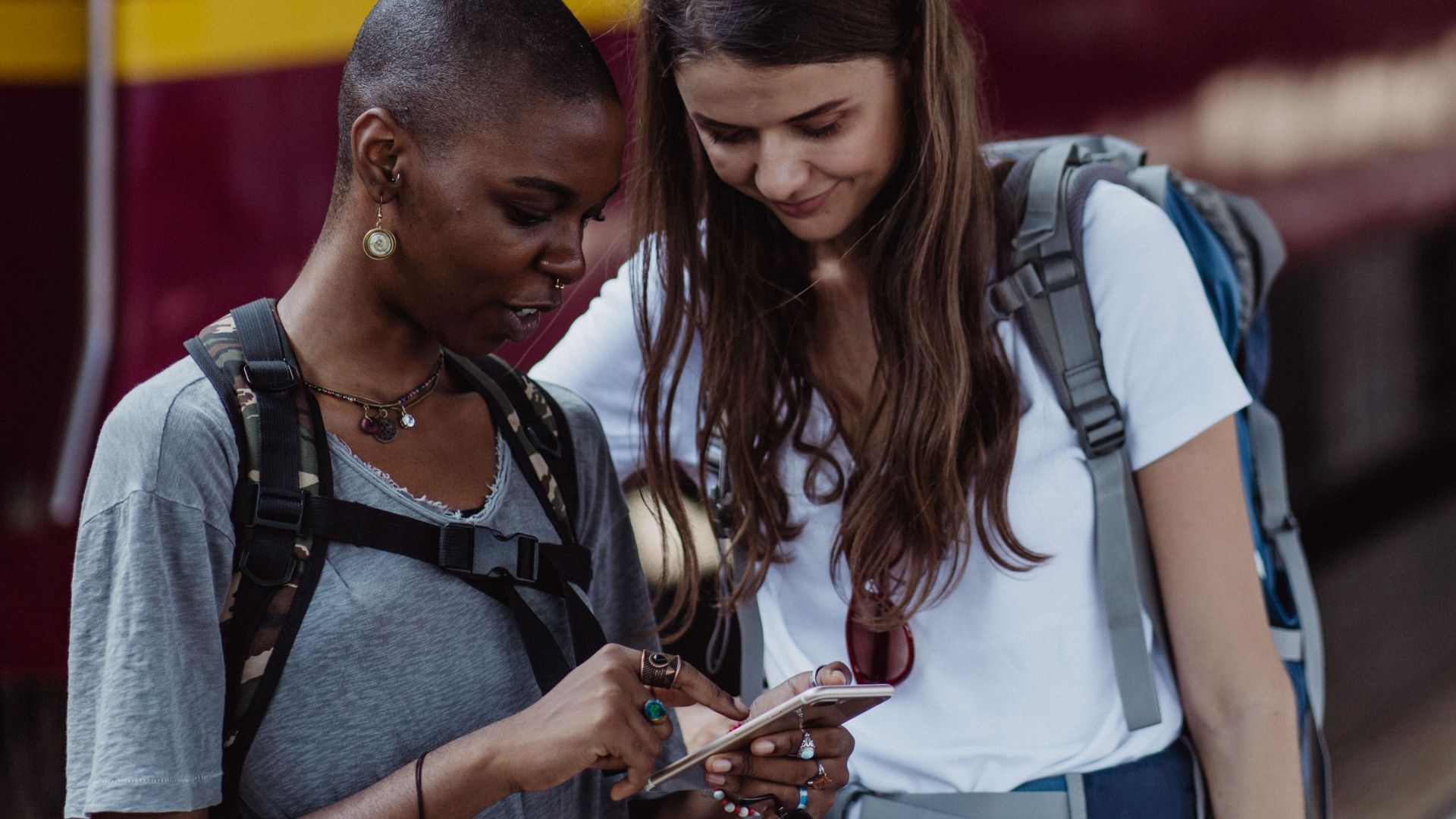 Two women backpackers checking phone at a train station, exploring travel options.