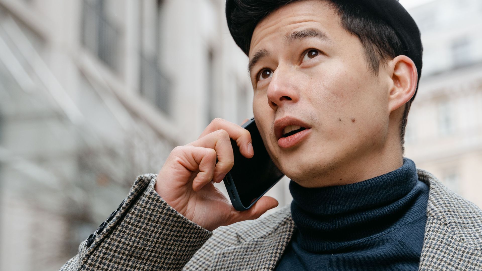 Stylish man in a city talking on a smartphone outdoors, wearing a coat and hat.