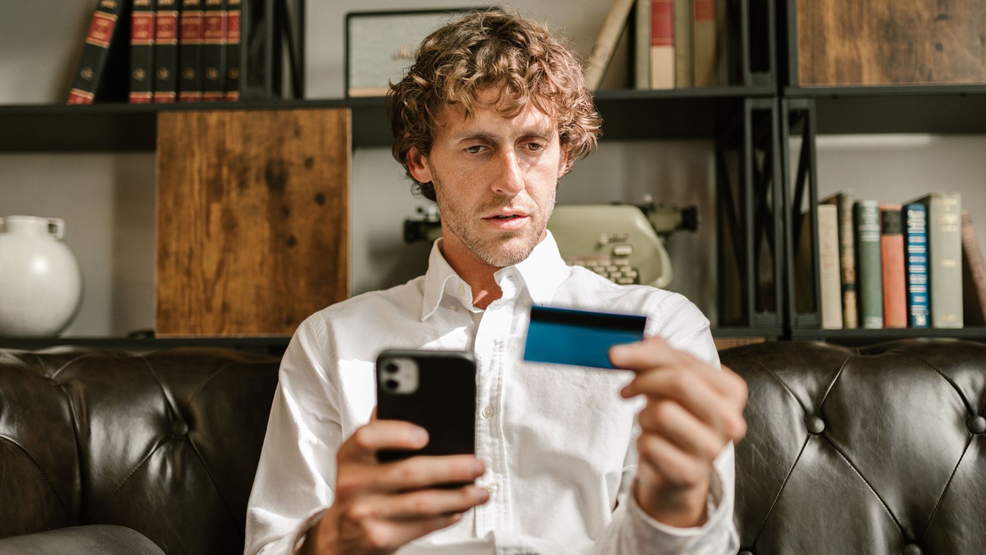 A man sitting on a leather sofa using a smartphone and credit card for online shopping.