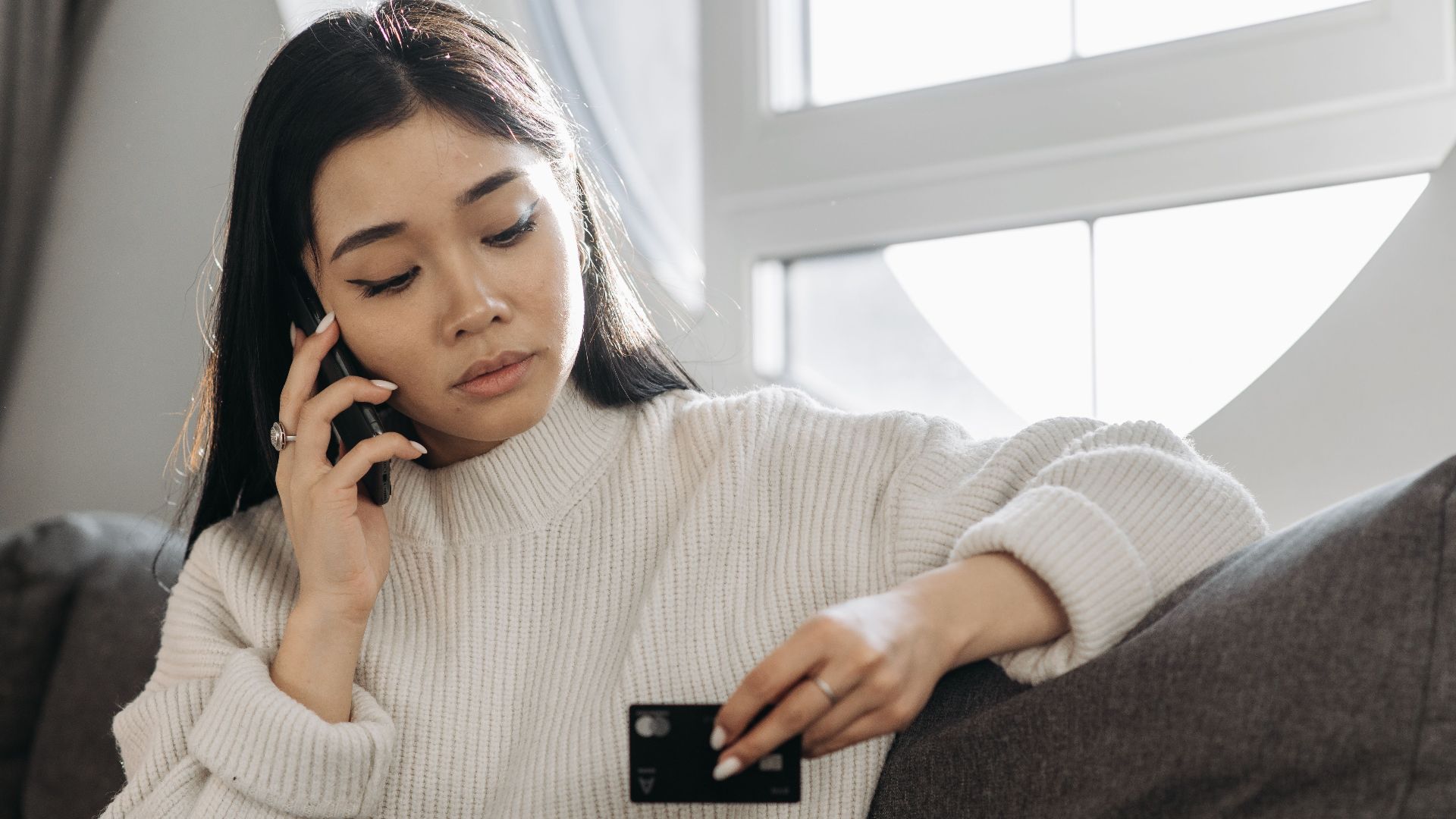 A woman sits on a sofa, multitasking with a phone and credit card, focusing on online shopping.