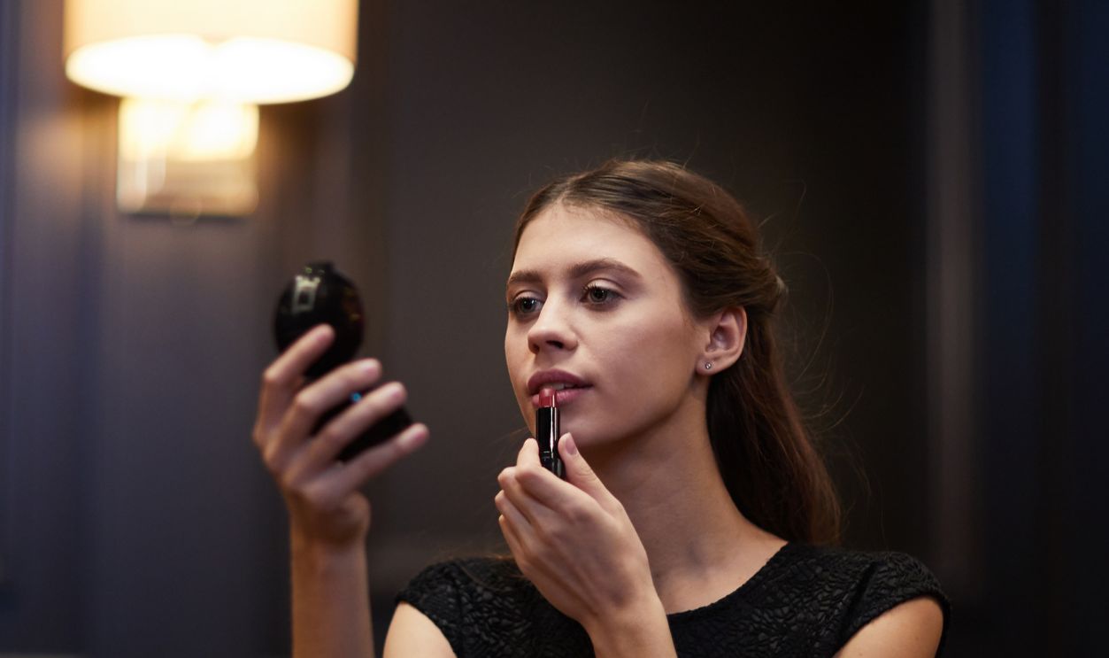 Young elegant woman applying lipstick on her lips while looking in compact powder mirror in restaurant
