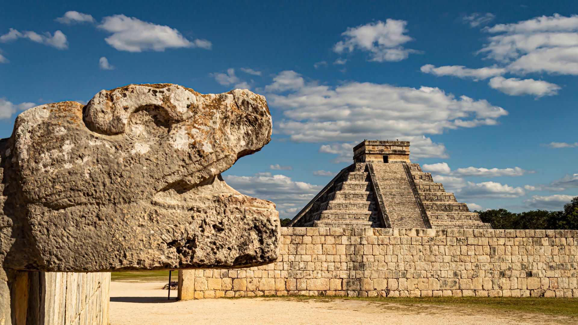 A sculpted stone head of Kukulkan (the Feathered Serpent) with the structure commonly known as the Castle (from Spanish El Castillo) in the background.