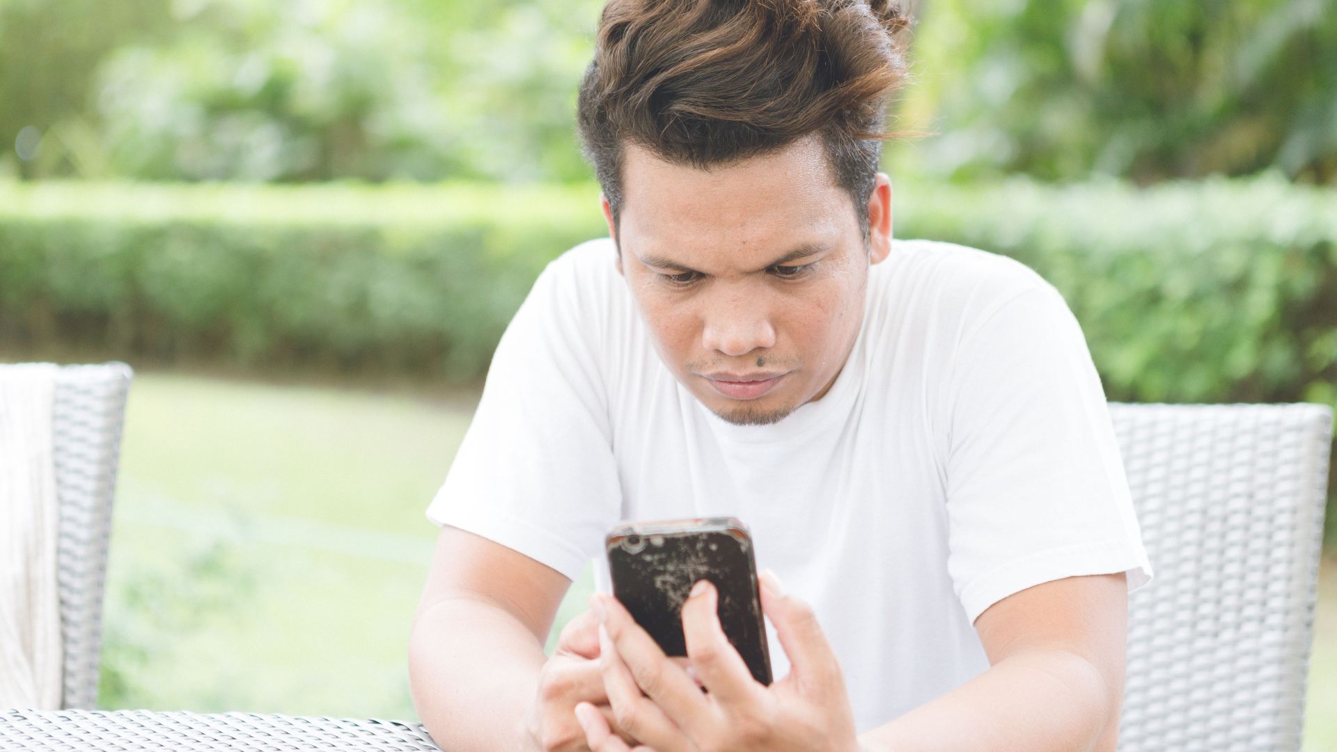 Concentrated ethnic male with stylish haircut wearing casual white shirt sitting on chair on sunny veranda and browsing cellphone