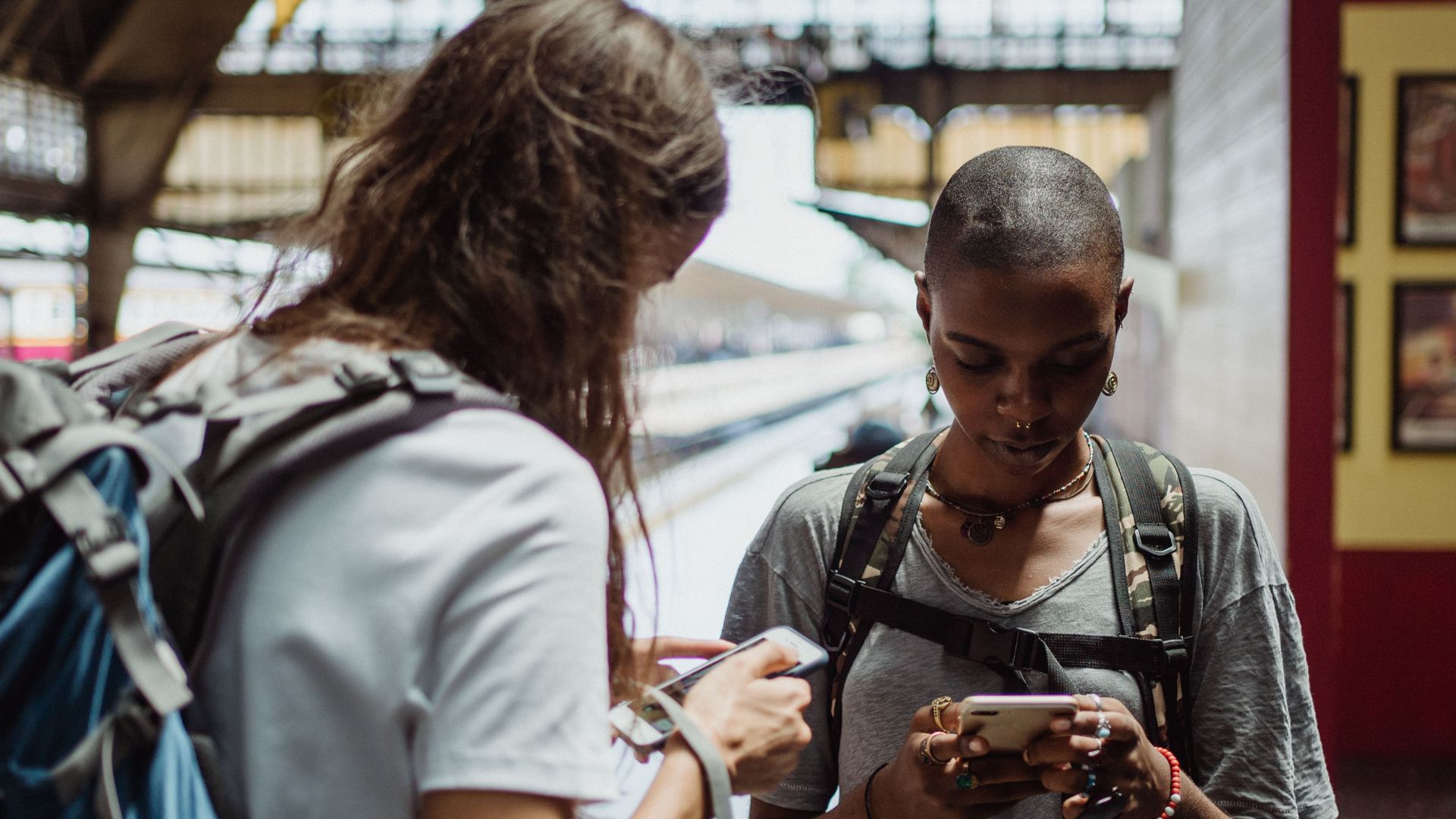Two travelers using smartphones while waiting at a railway station, backpacks on their shoulders.