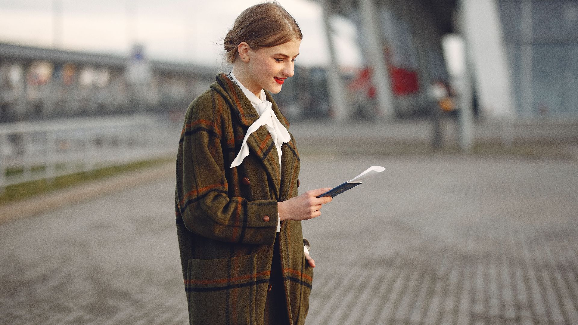 Smiling female passenger wearing trendy plaid coat and white blouse checking passport and ticket standing on pavement near contemporary building of airport