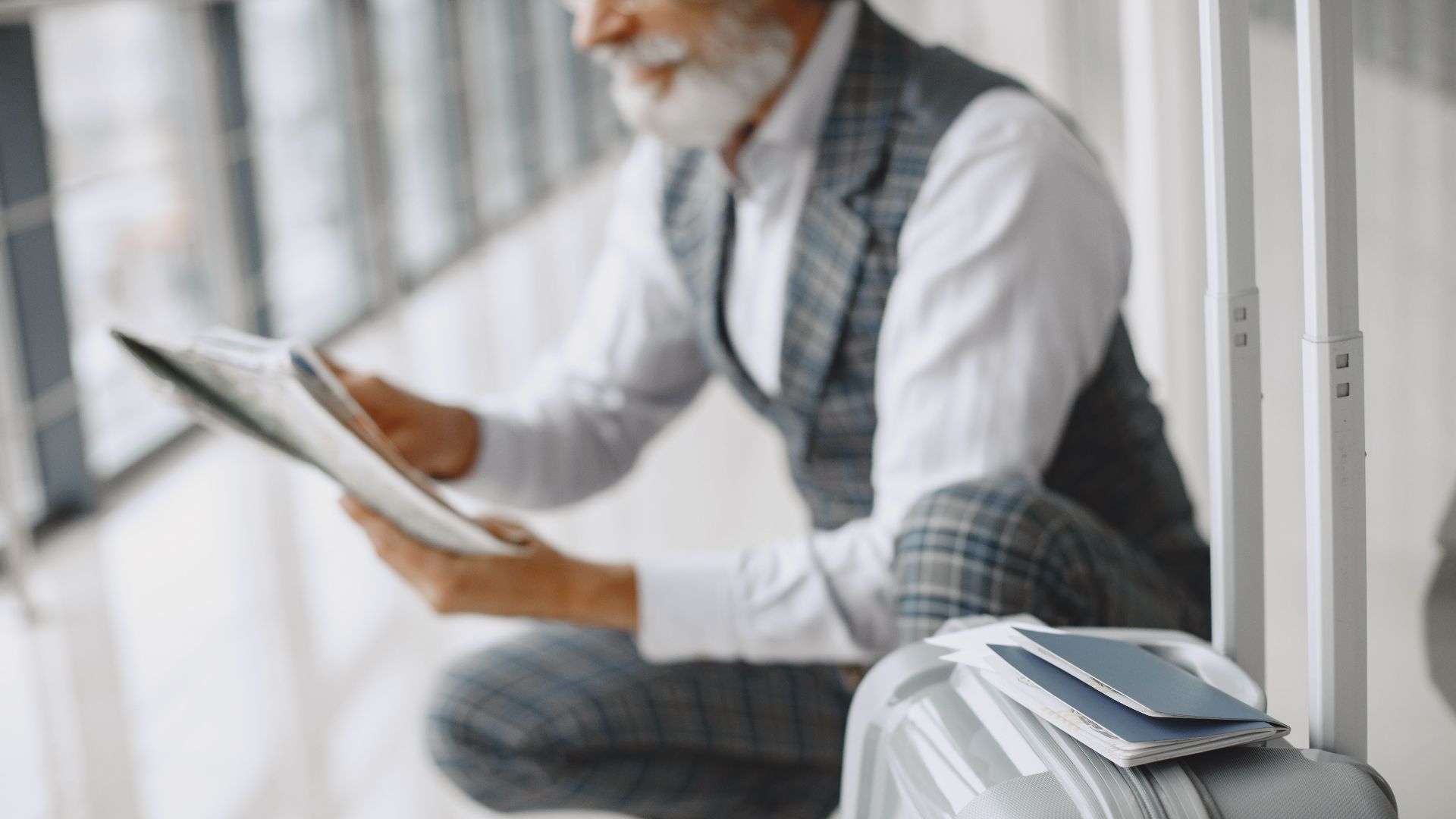 Adult man in plaid suit reads newspaper with suitcase at airport terminal.