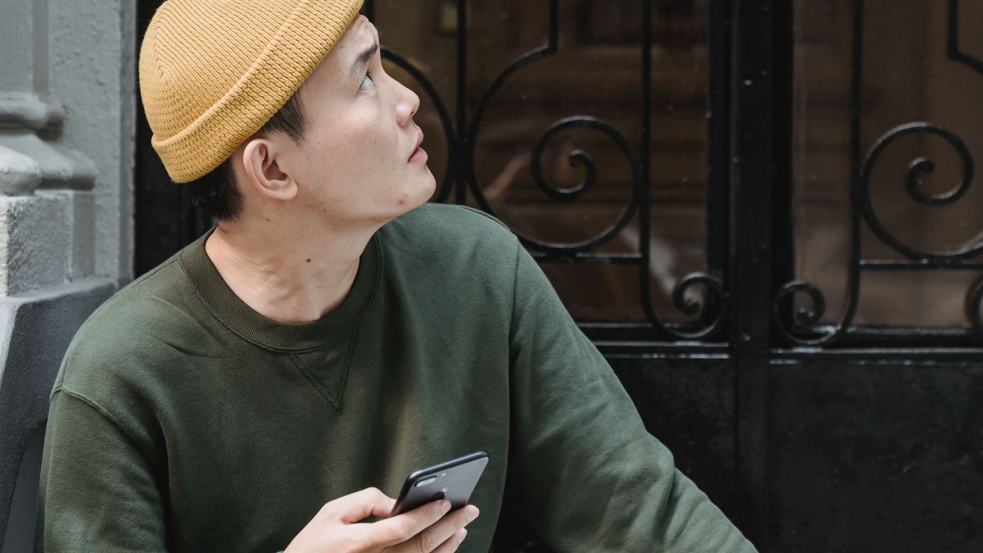 A puzzled delivery man sits outdoors, holding a phone and box, looking up at a door.