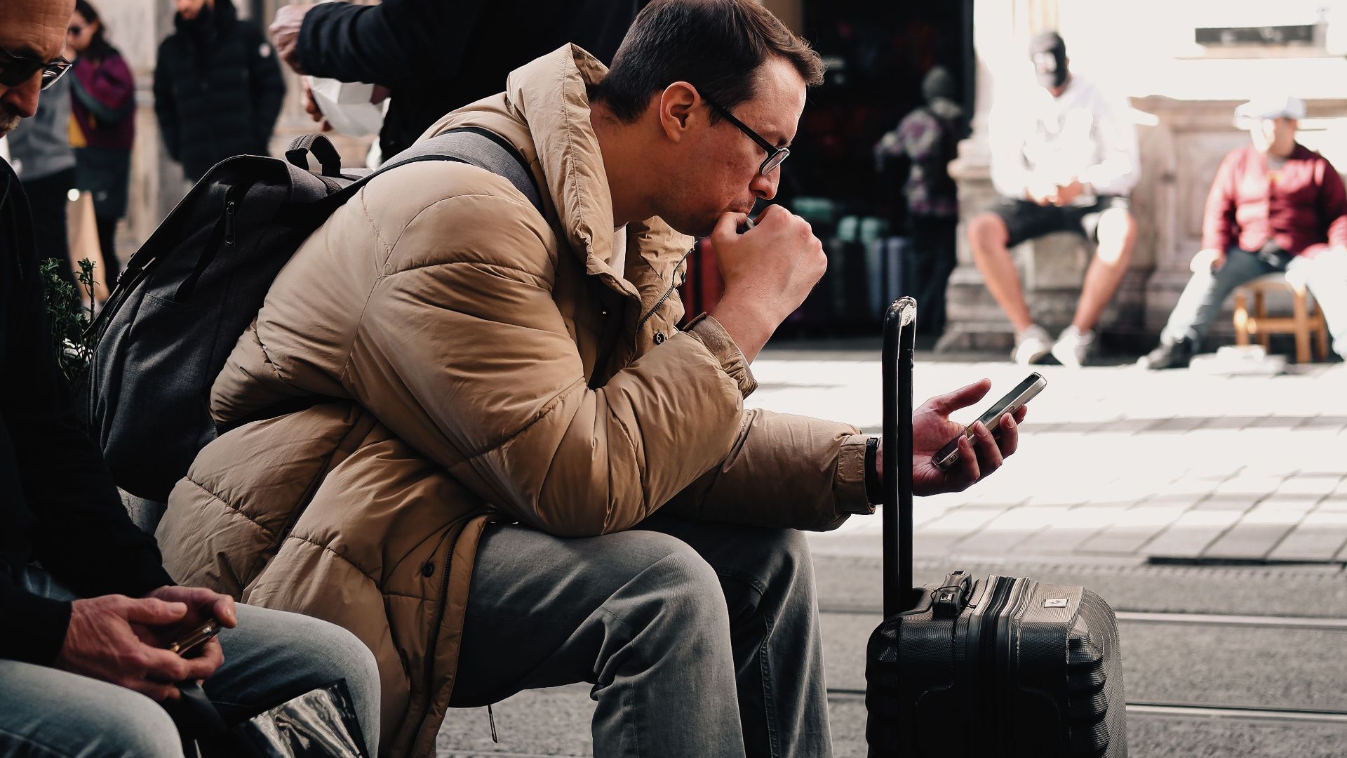 A man sitting with luggage, checking his phone in a bustling urban street scene, surrounded by people.