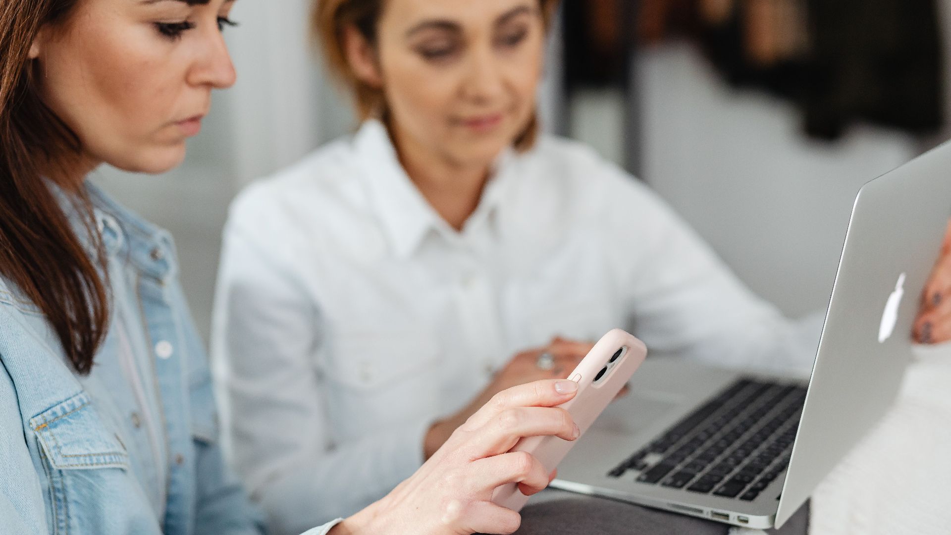 Two women working together using a laptop and smartphone in a casual setting.