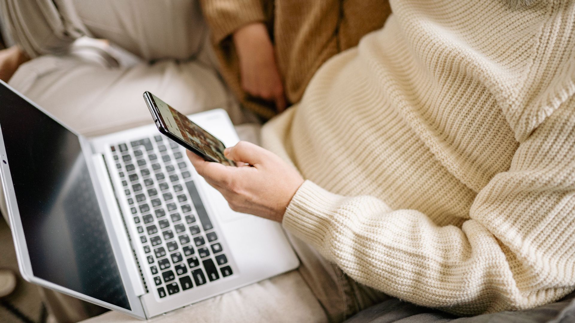 Adult in a cozy sweater multitasking with a smartphone and laptop indoors.