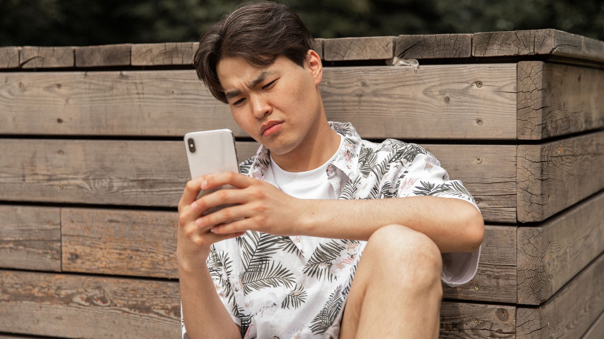 Young ethnic male in stylish clothes leaning on wooden barrier while messaging on mobile phone in park