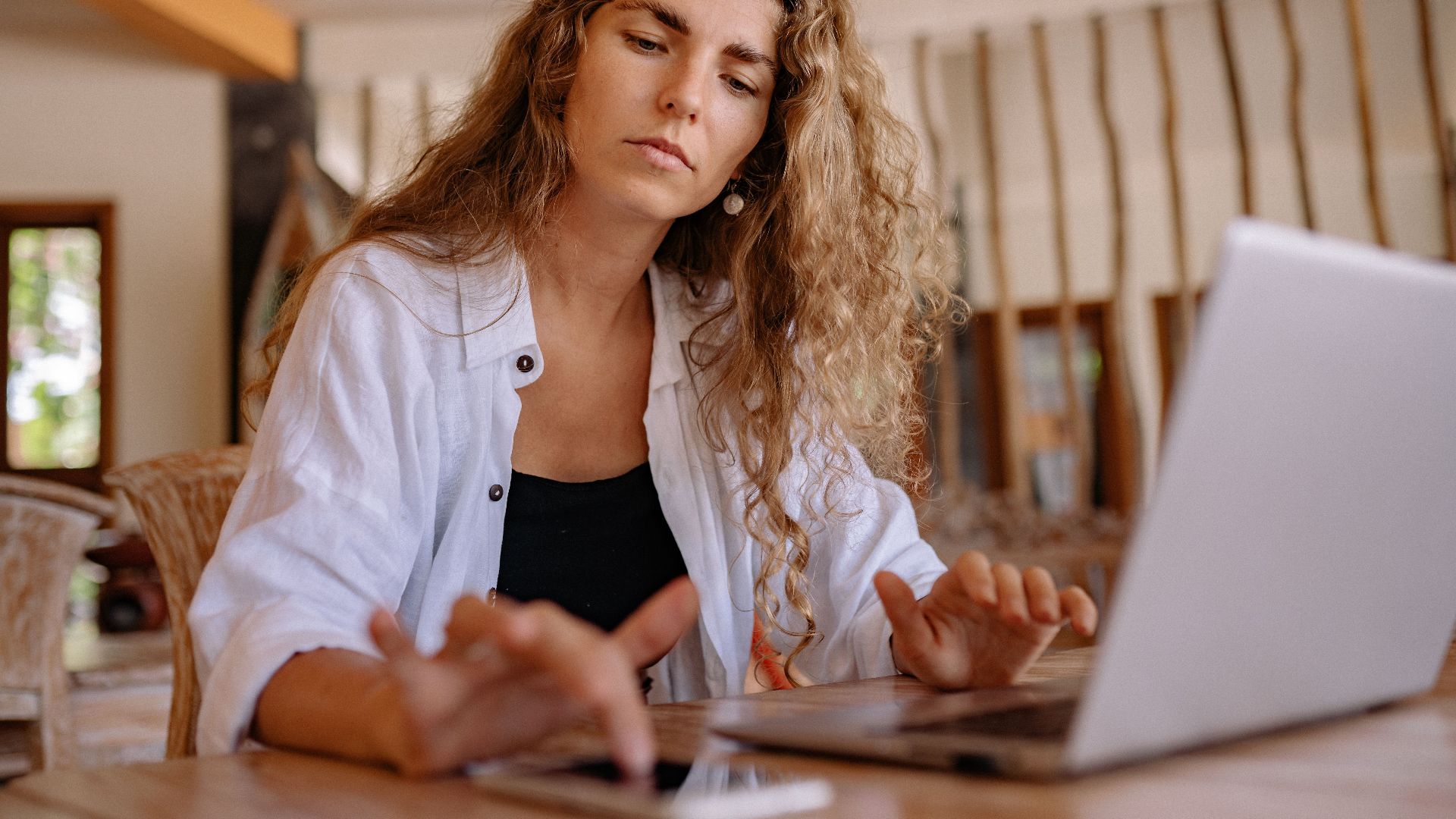 A woman intently works on a laptop while using a smartphone indoors, symbolizing modern remote work.