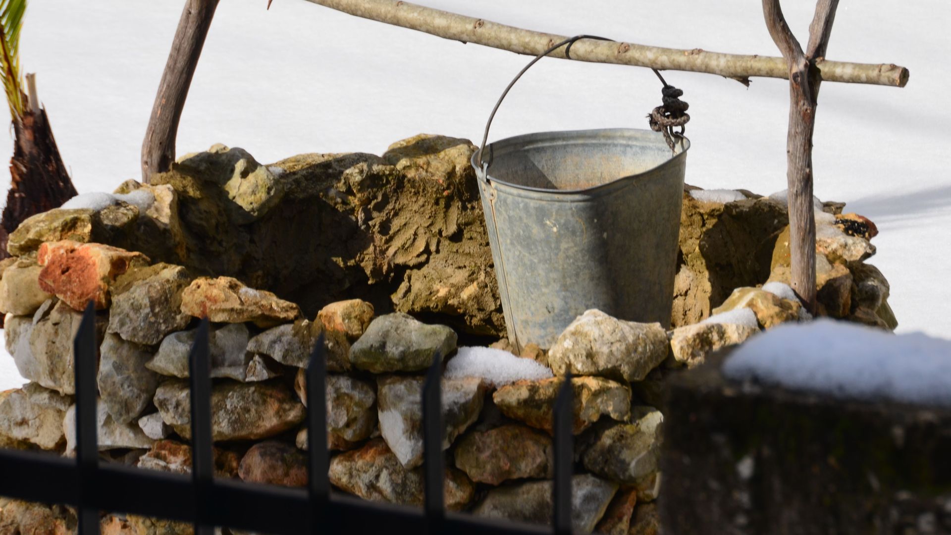 A rustic stone well with a metal bucket and wooden branches in an outdoor setting.