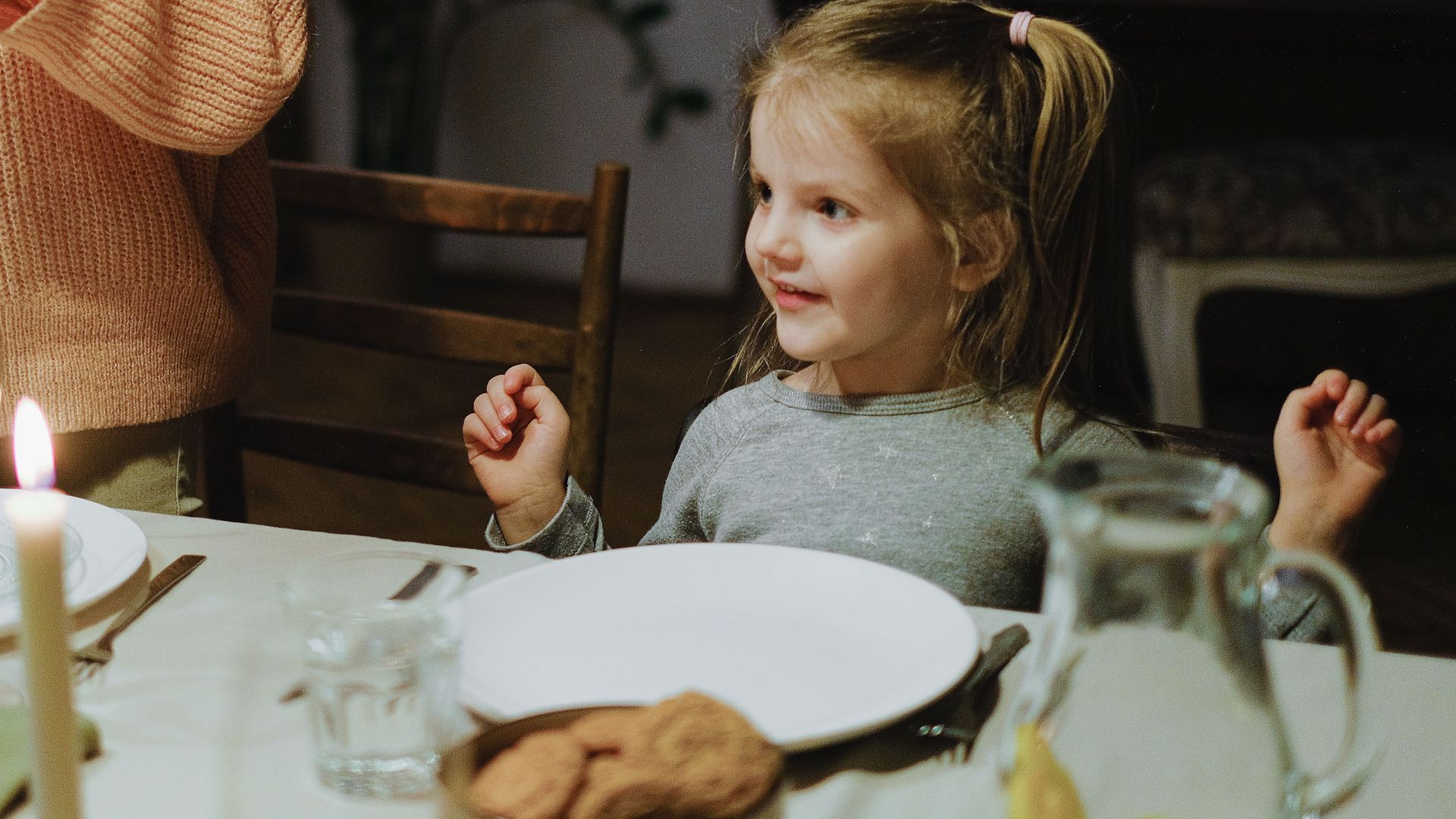 A young girl eagerly awaits a meal at a candlelit family dinner table indoors.